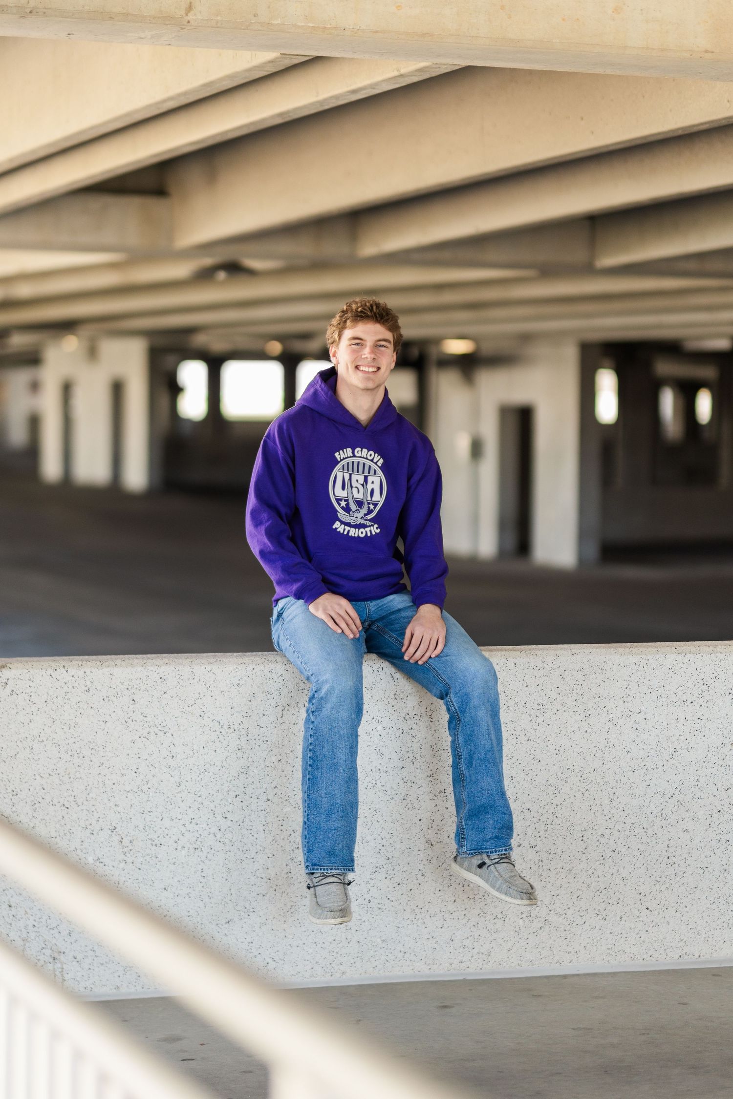 Someone in a purple university sweatshirt sits on a concrete barrier in a parking garage.