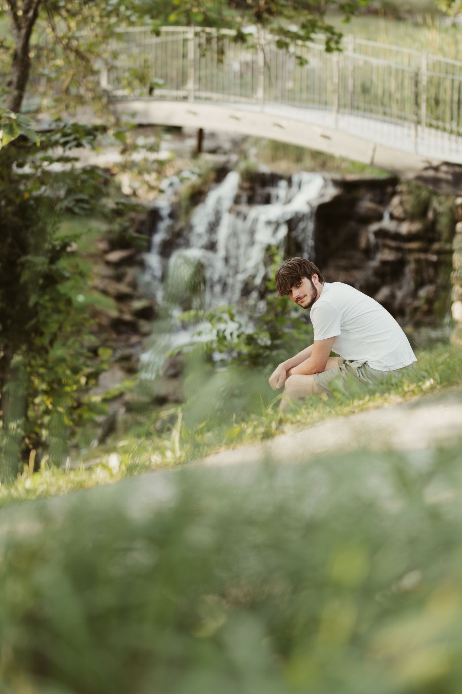 A person in white crouches near a scenic waterfall with a bridge visible in the background at a peaceful park.