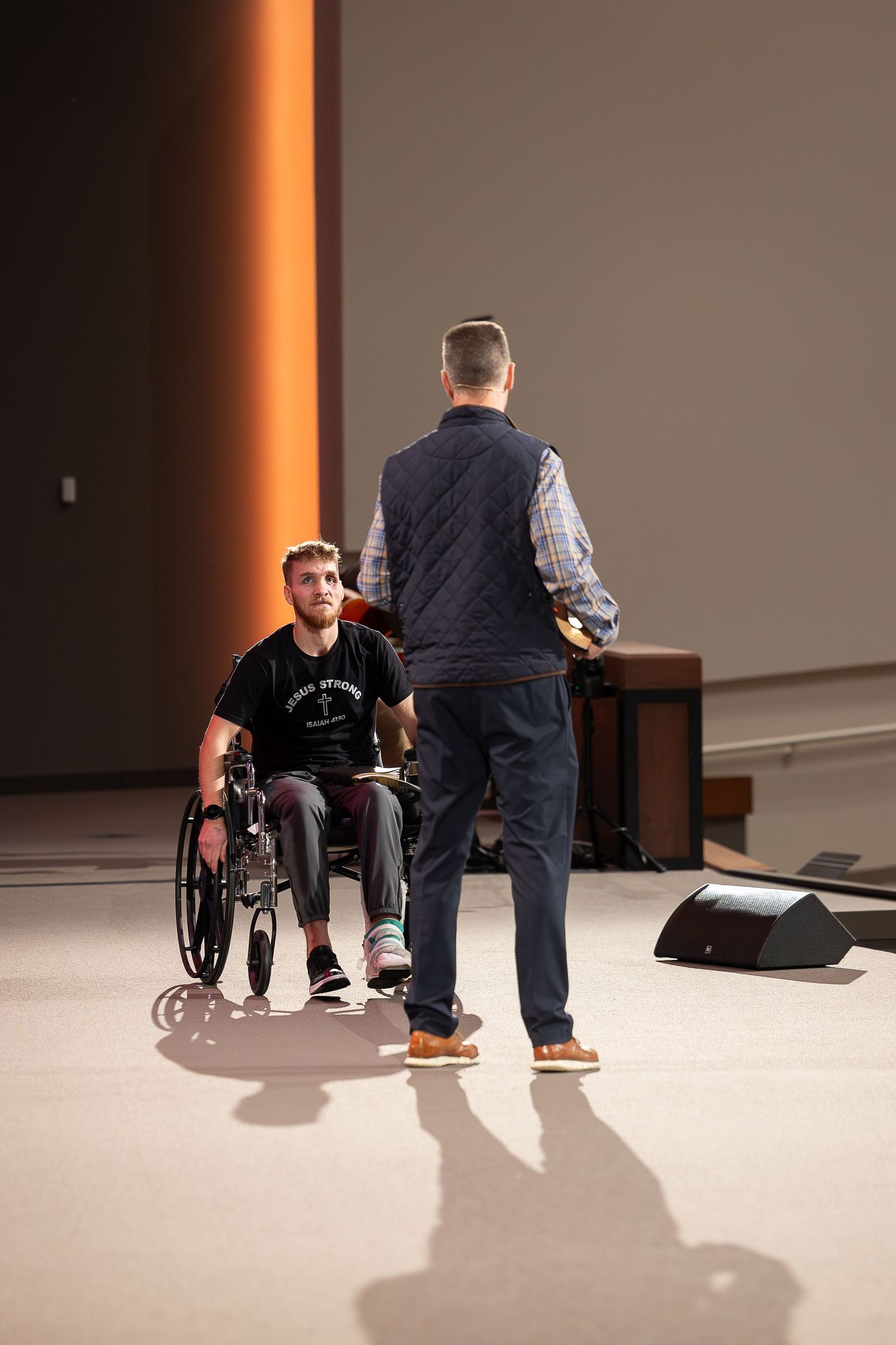 Two individuals stand in conversation on an indoor stage, with one person seated in a wheelchair and another standing.