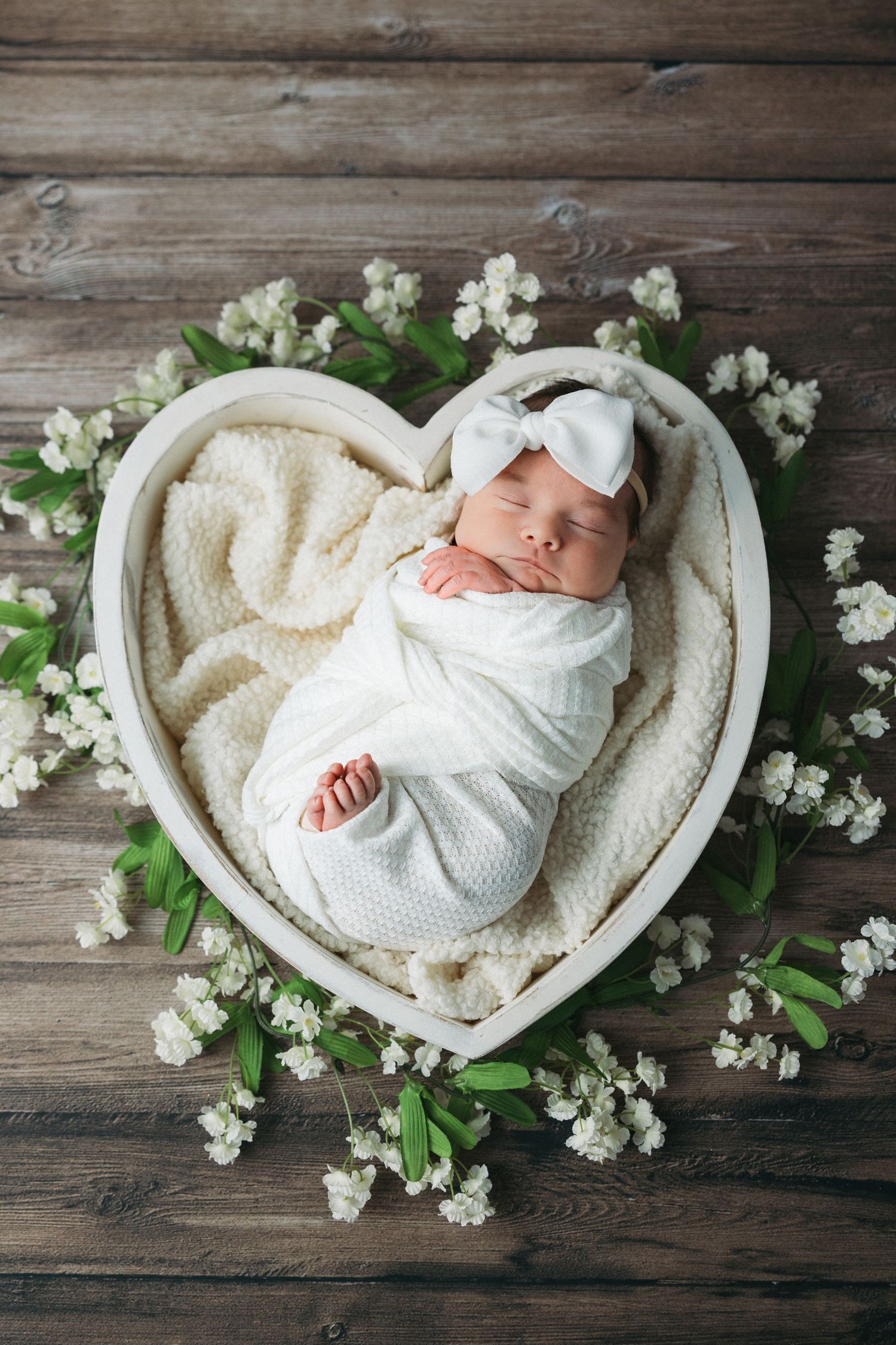 Sleeping newborn swaddled in white blanket lies in heart-shaped basket surrounded by delicate white flowers on wooden surface.