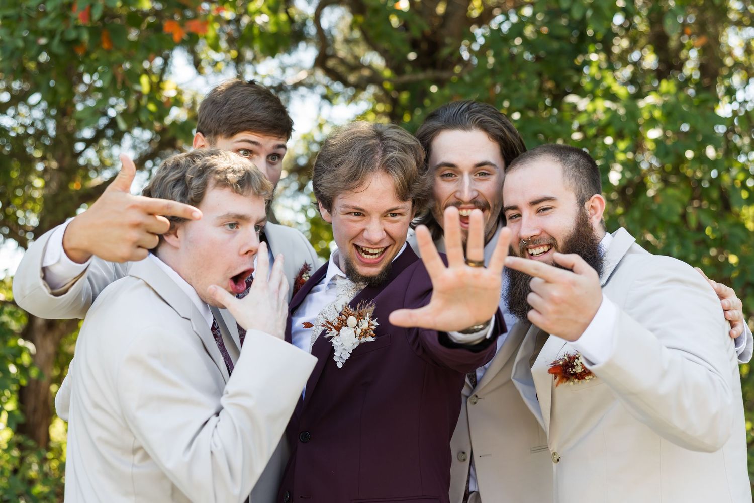 Groom shows off wedding ring surrounded by groomsmen making fun poses outdoors at wedding celebration.