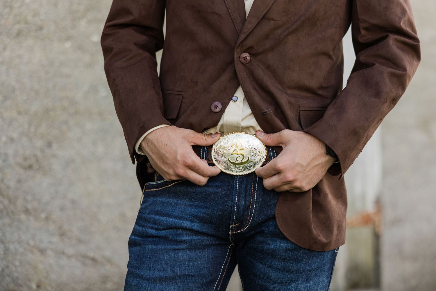 Man wearing brown blazer and jeans with a large decorative Western silver belt buckle.