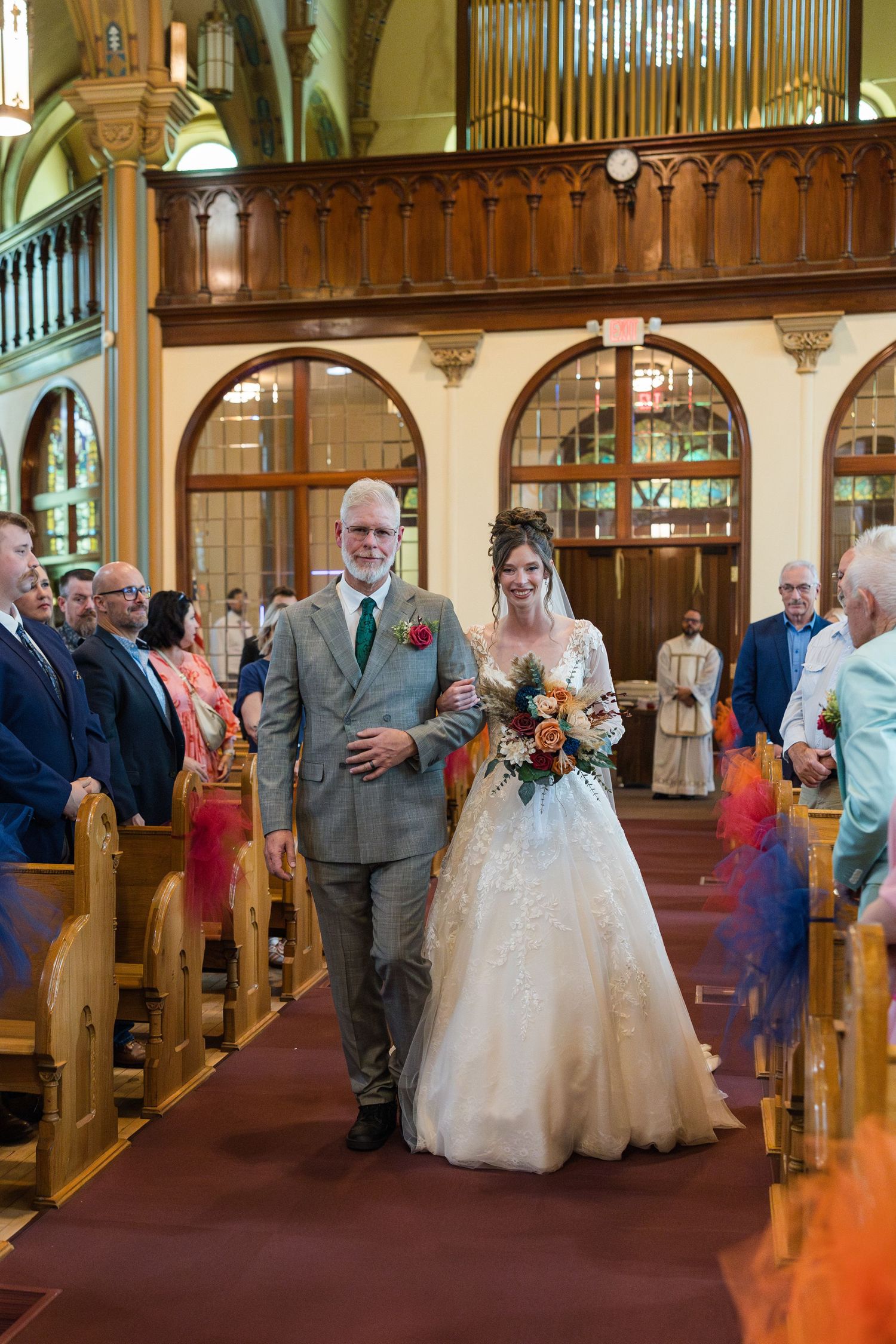 Bride walks down church aisle with escort at wedding ceremony, guests seated in wooden pews.
