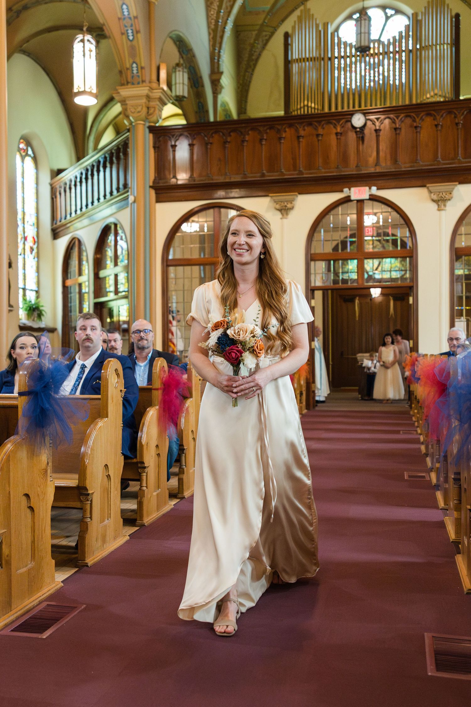Smiling bridesmaid in gold satin dress walks down church aisle carrying colorful bouquet at wedding ceremony.