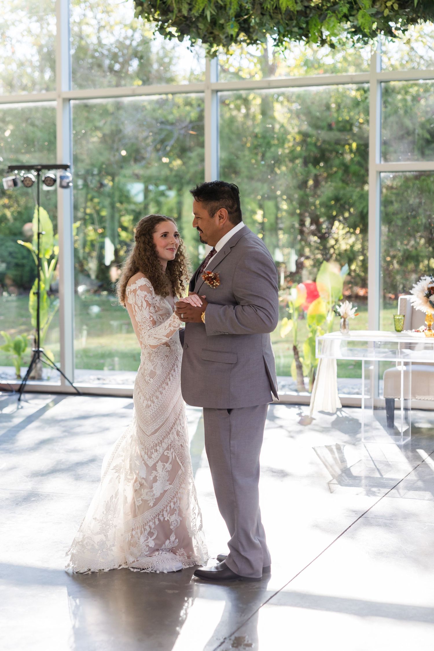 Bride and groom share their first dance in a sunlit glass venue surrounded by lush outdoor scenery.