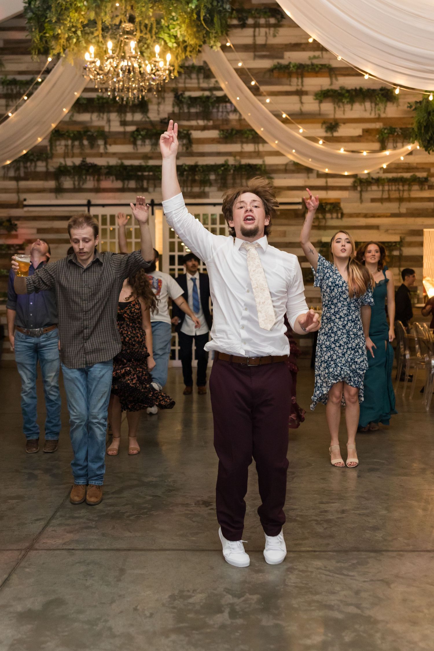 Energetic wedding guest in white shirt and maroon pants dances with arms raised at rustic barn reception.