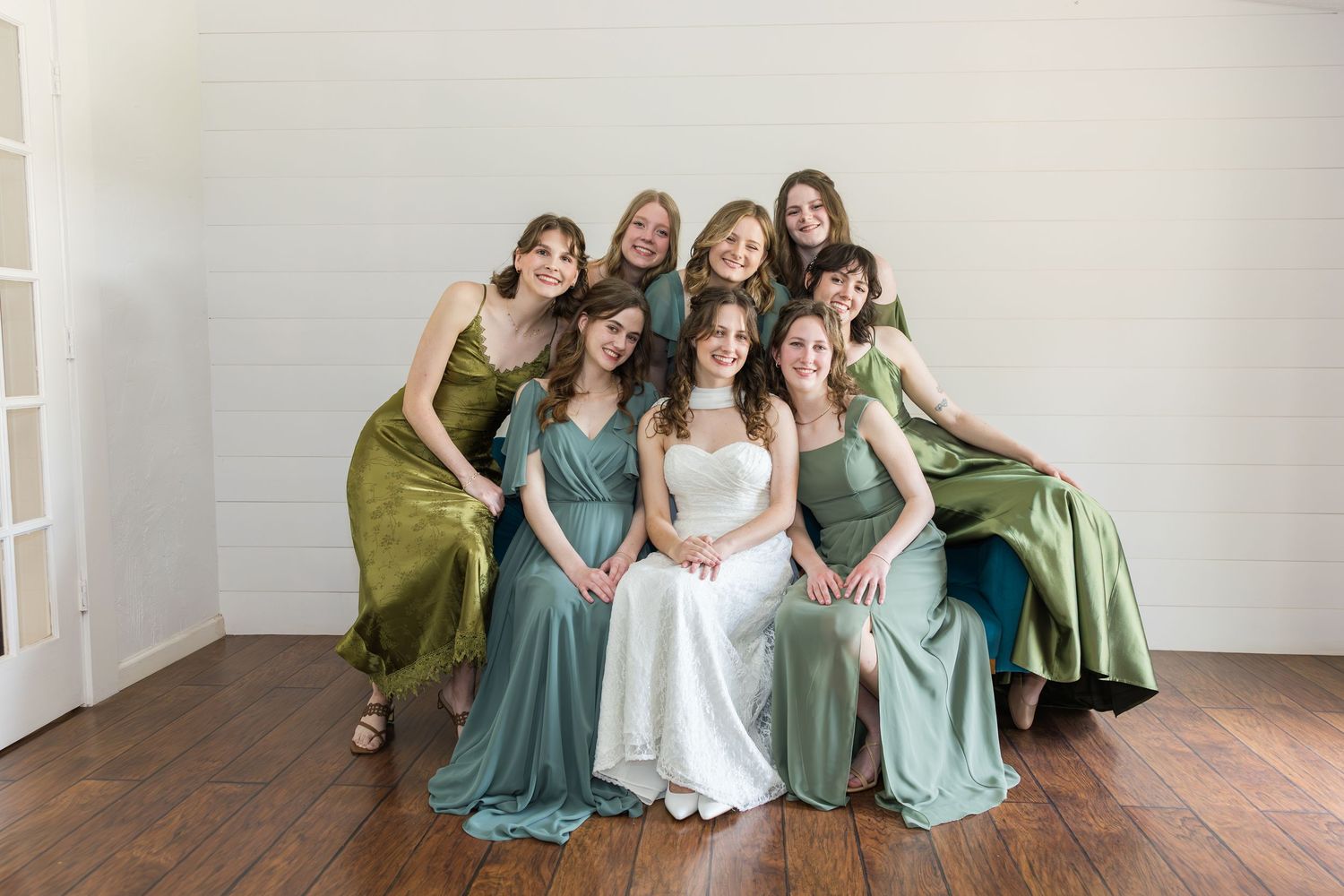 Bride poses with seven bridesmaids in elegant green satin dresses inside a bright white venue.