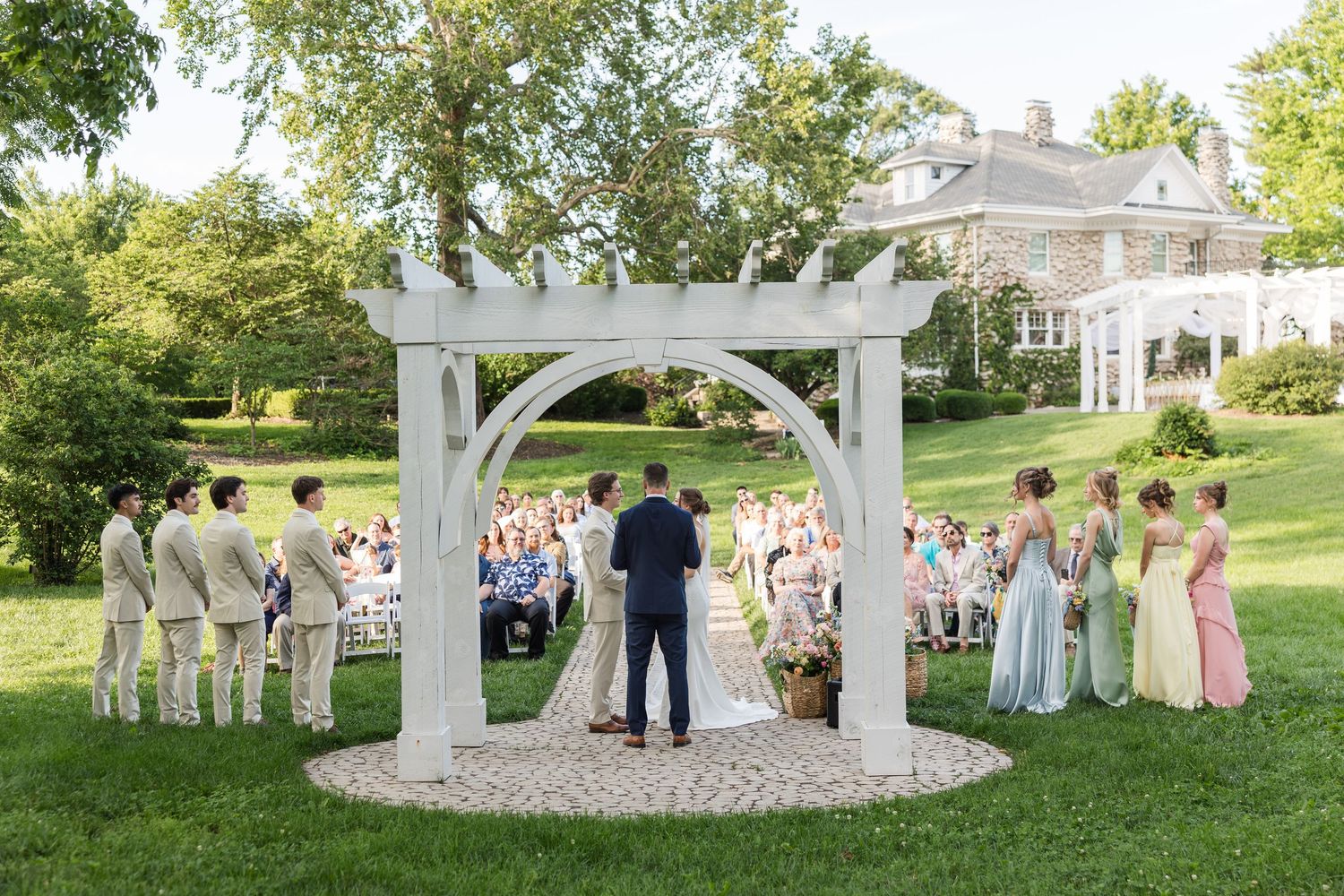 Outdoor wedding ceremony under white arch with bridal party on green lawn beside elegant estate.