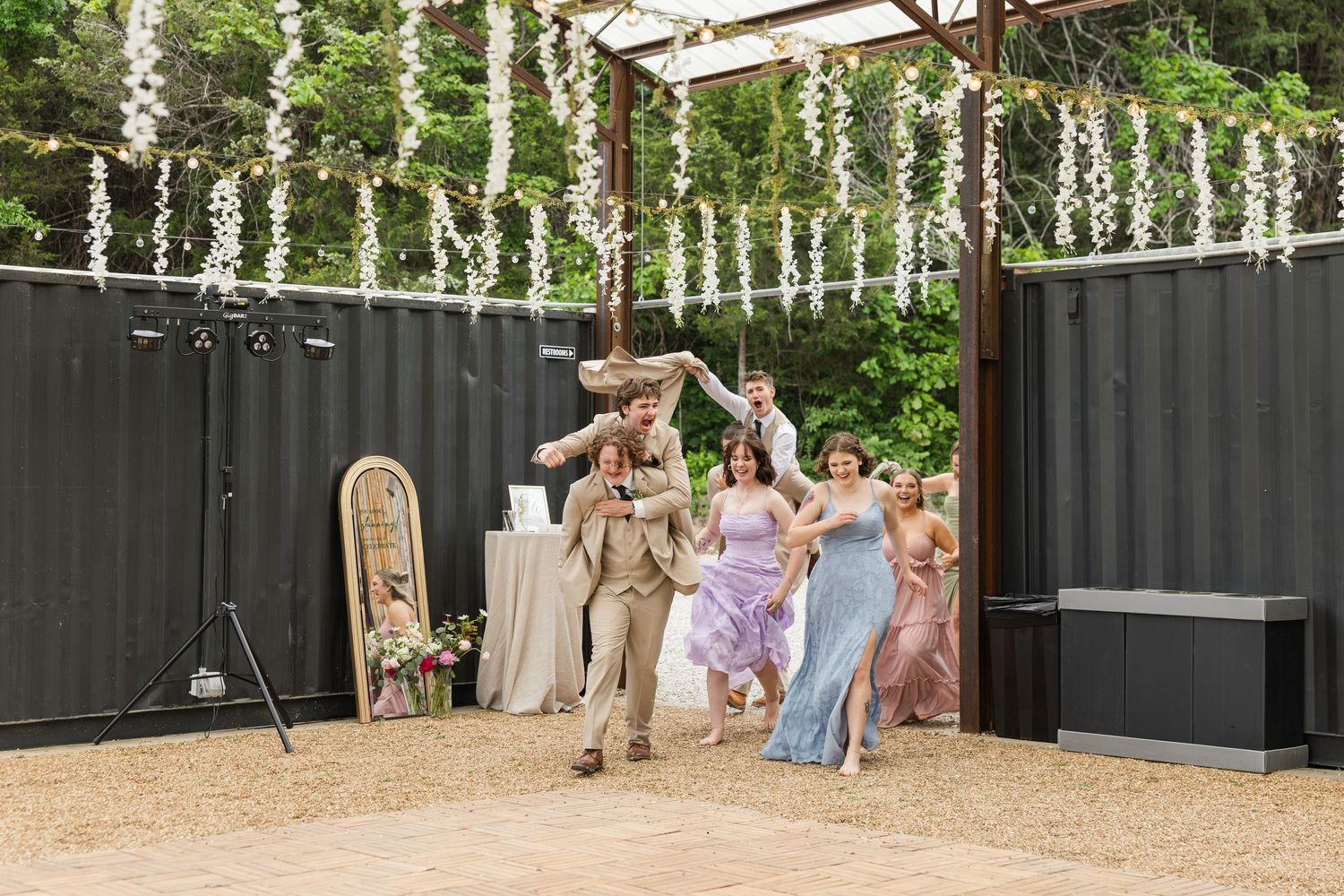 Wedding party celebrating outdoors under floral garland pergola, with groomsmen lifting groom joyfully.