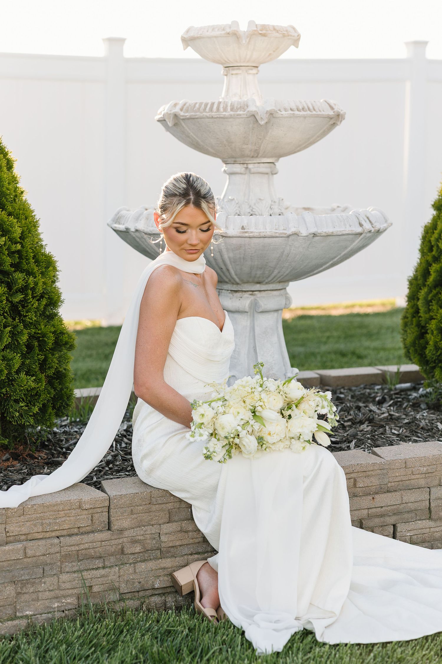Elegant bridal portrait with white gown and bouquet against ornate garden fountain.