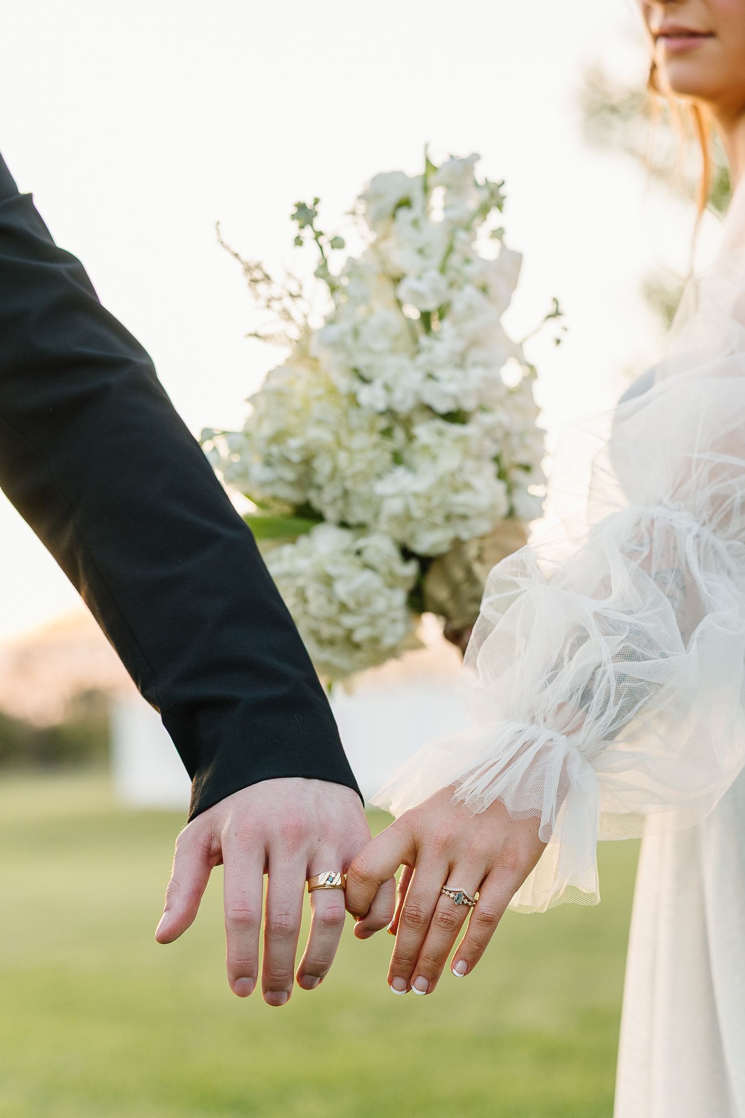 Close-up view of newlywed couple showing off their wedding rings with white floral decor in the background.