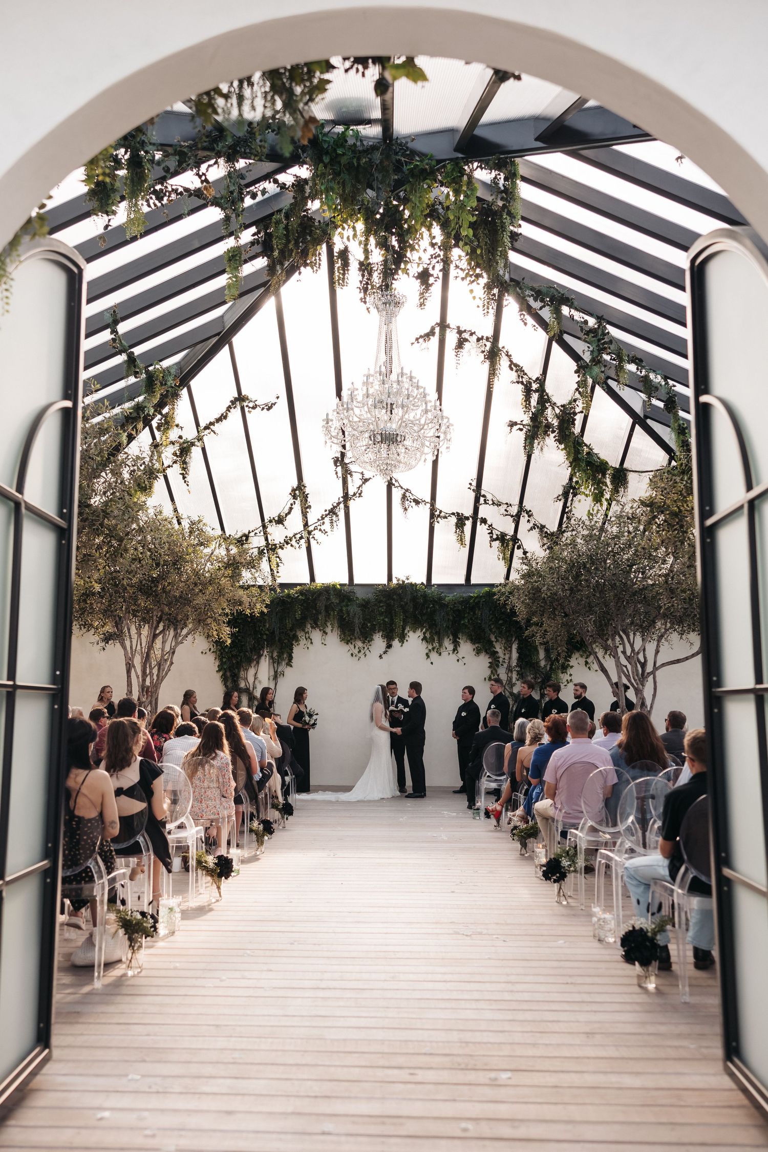 Wedding ceremony in a modern white chapel with glass ceiling, hanging greenery and crystal chandelier.