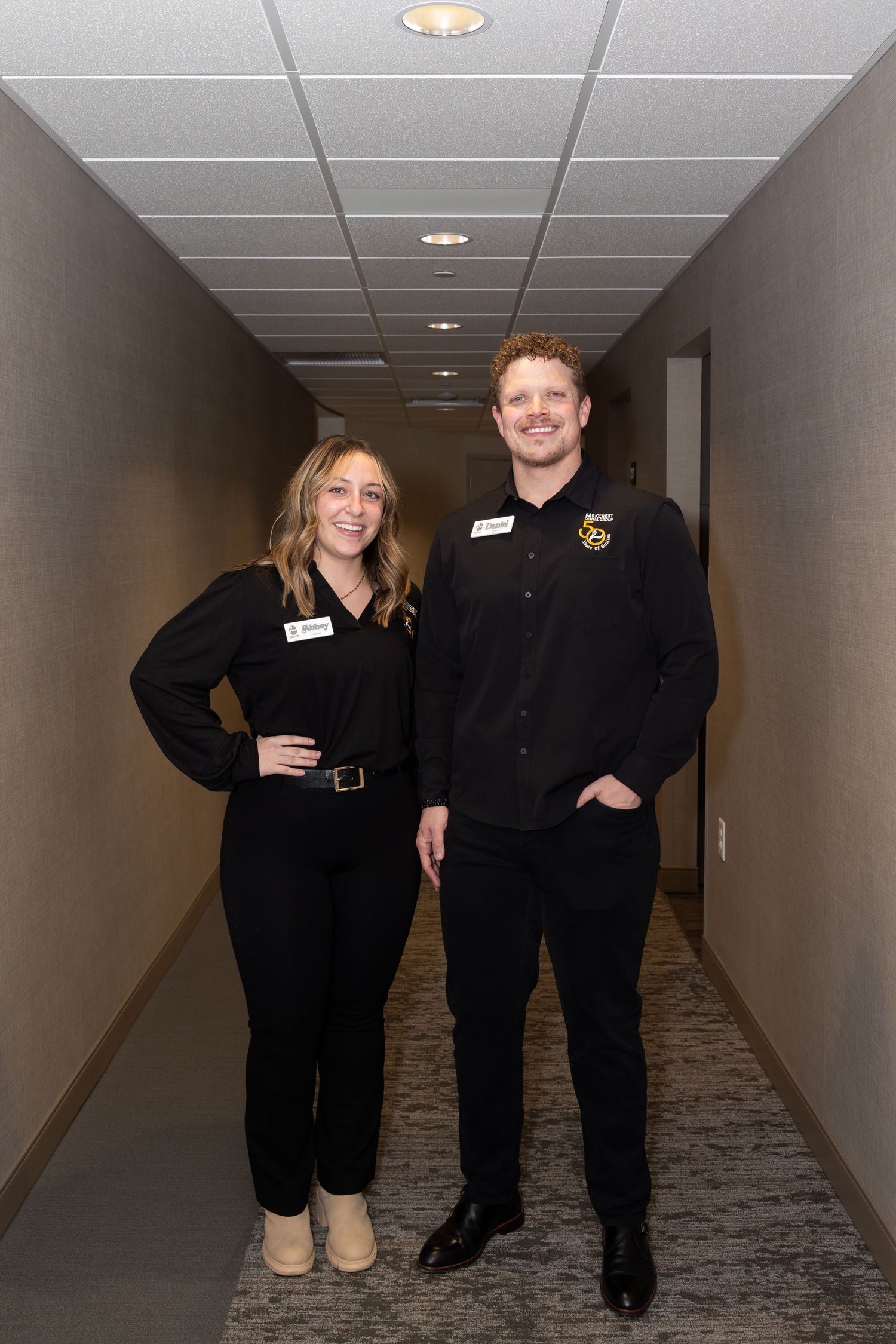 Two staff members in black uniforms standing in a carpeted office hallway.