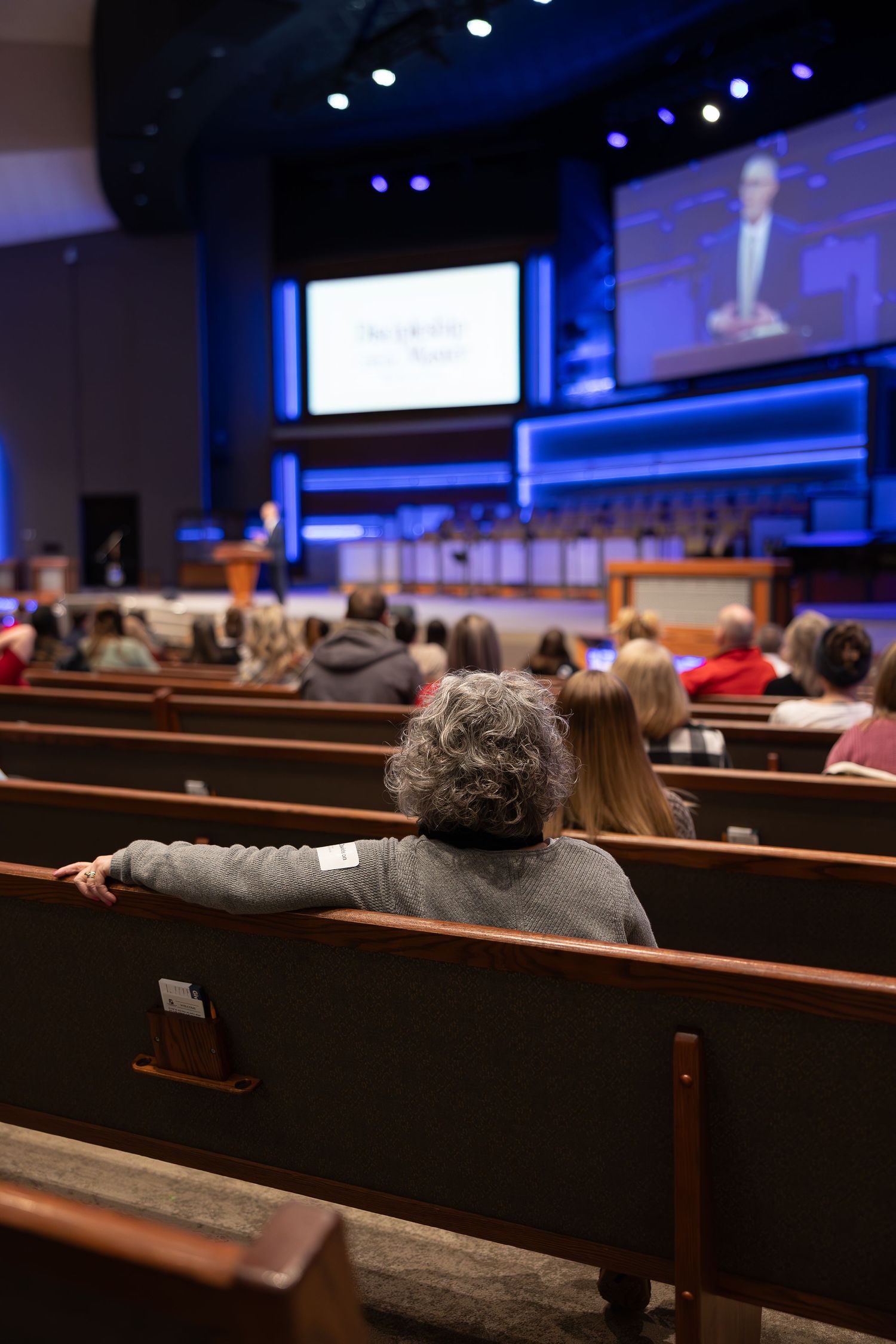 View from behind of churchgoers seated in wooden pews during a service with screens visible.