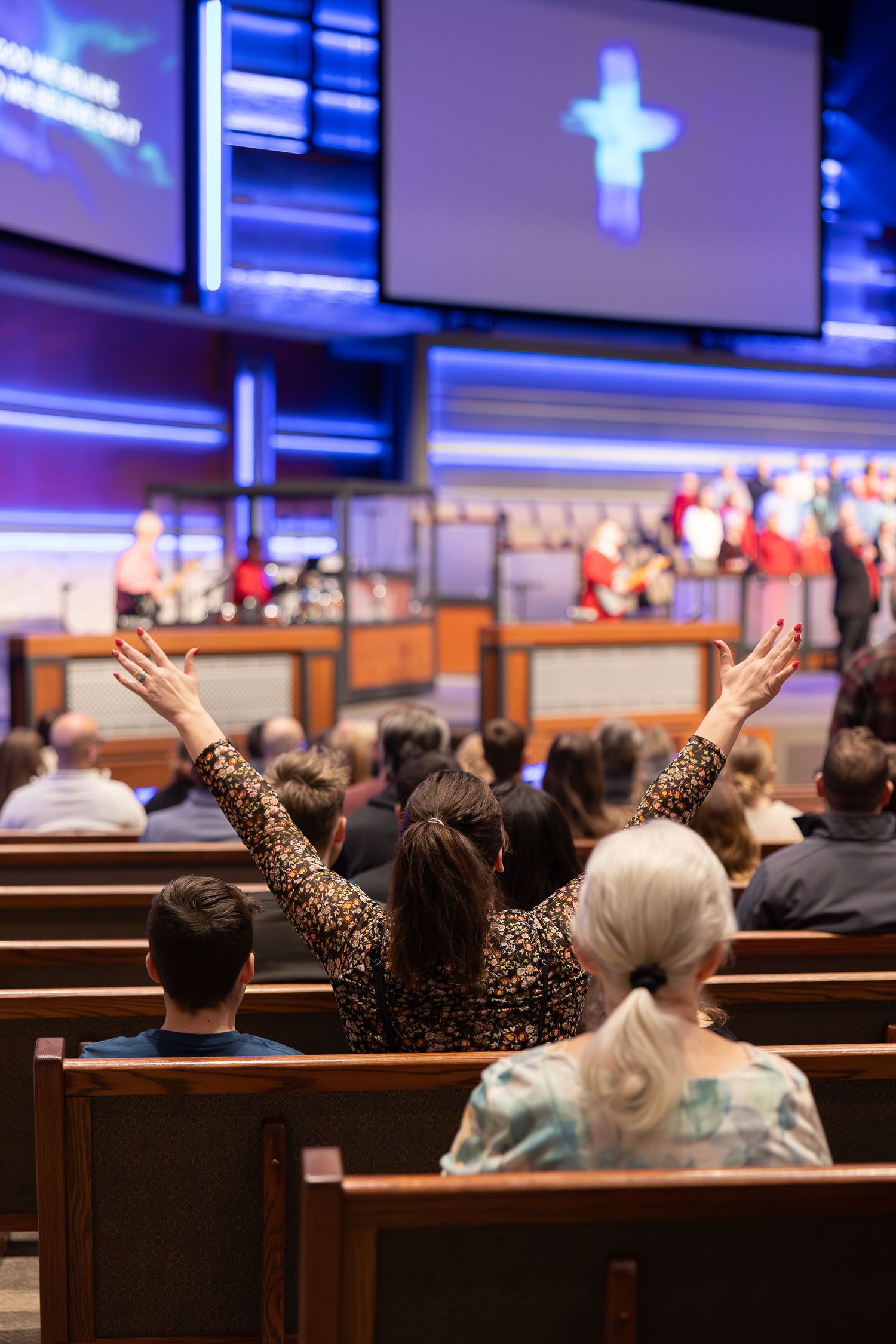 People worshipping in church pews with raised hands and illuminated cross on screen.