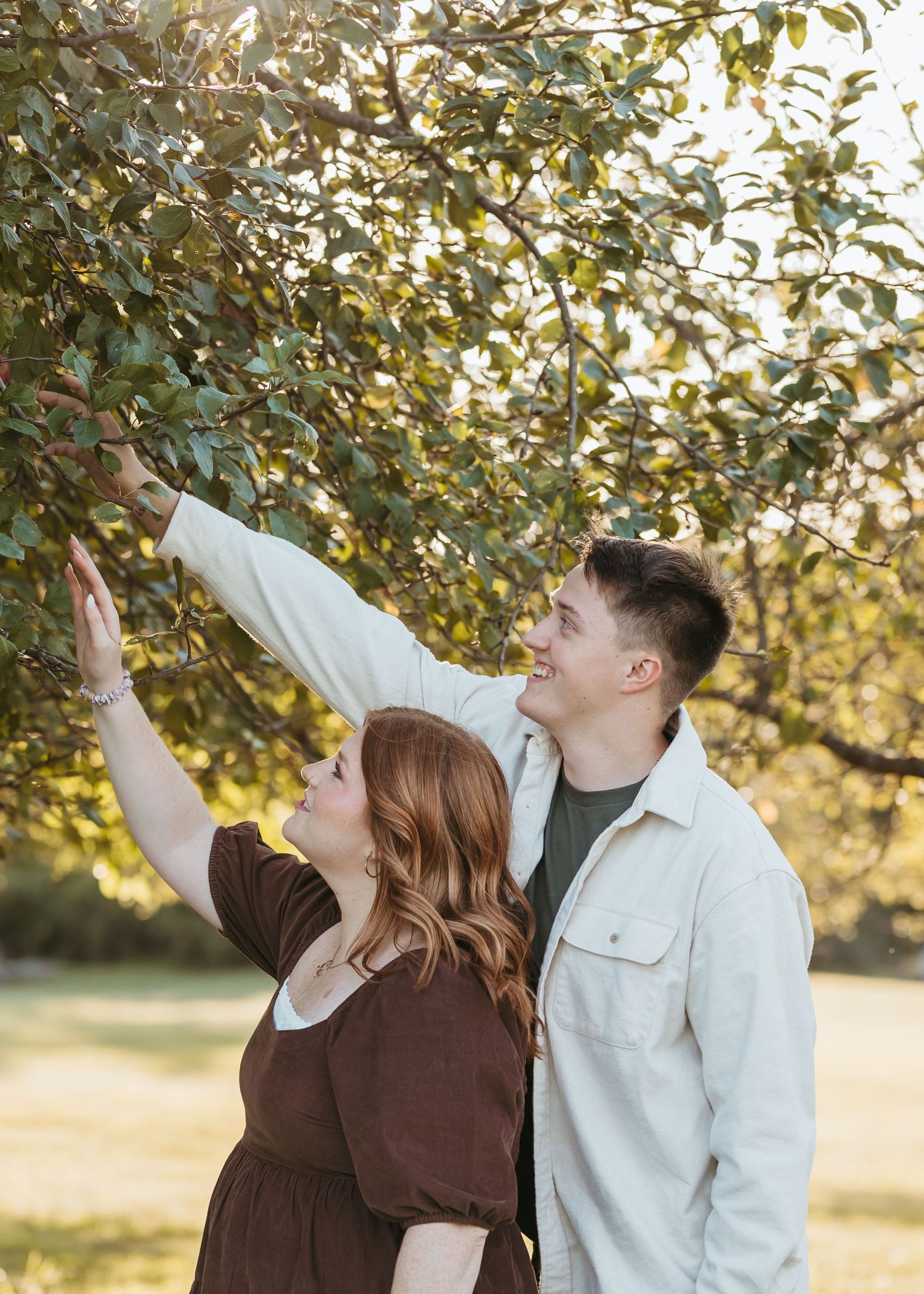Two people reach up toward autumn leaves in a sunlit outdoor setting.