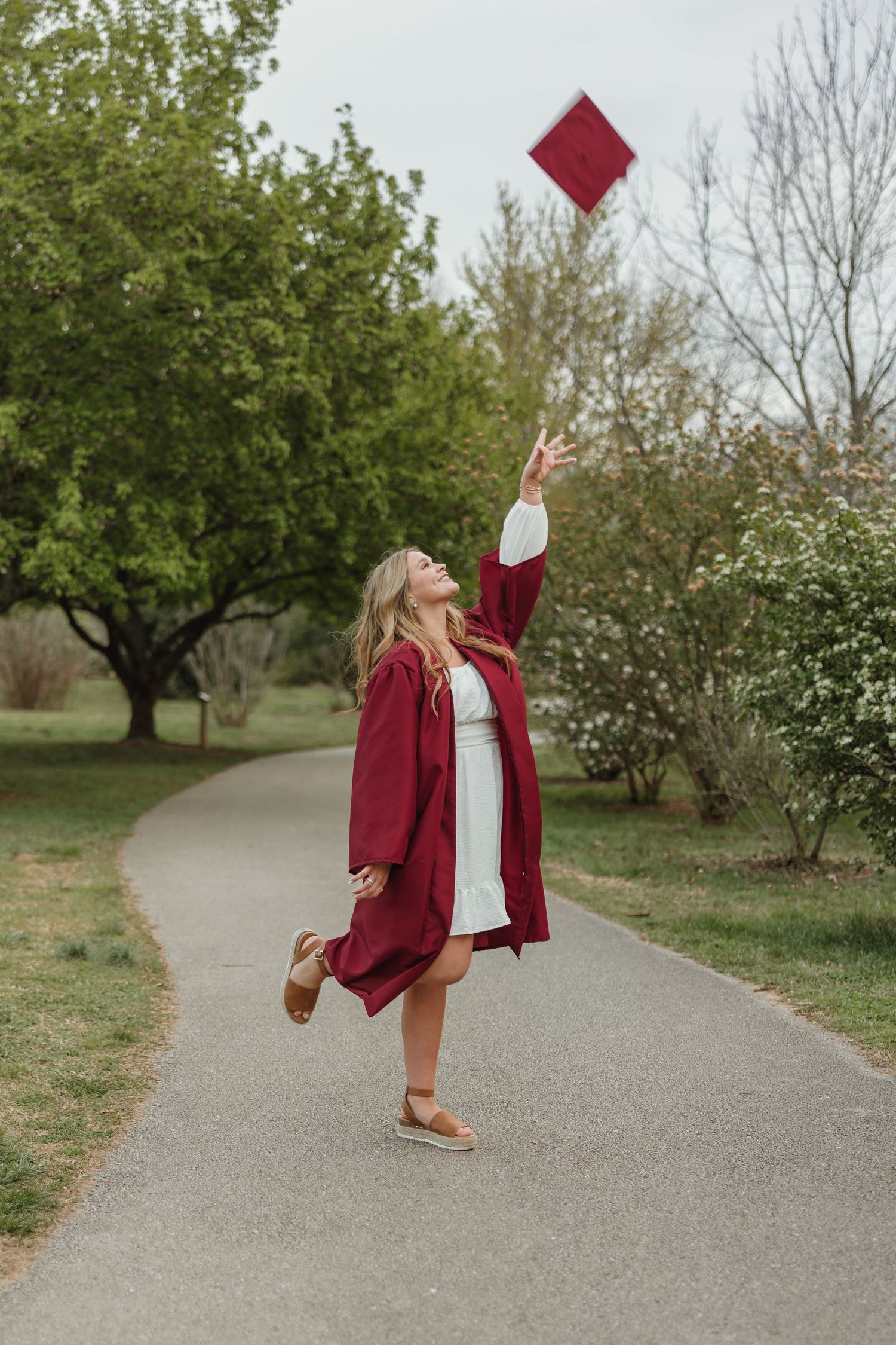 Graduate in maroon gown tosses cap in celebratory pose on garden path.