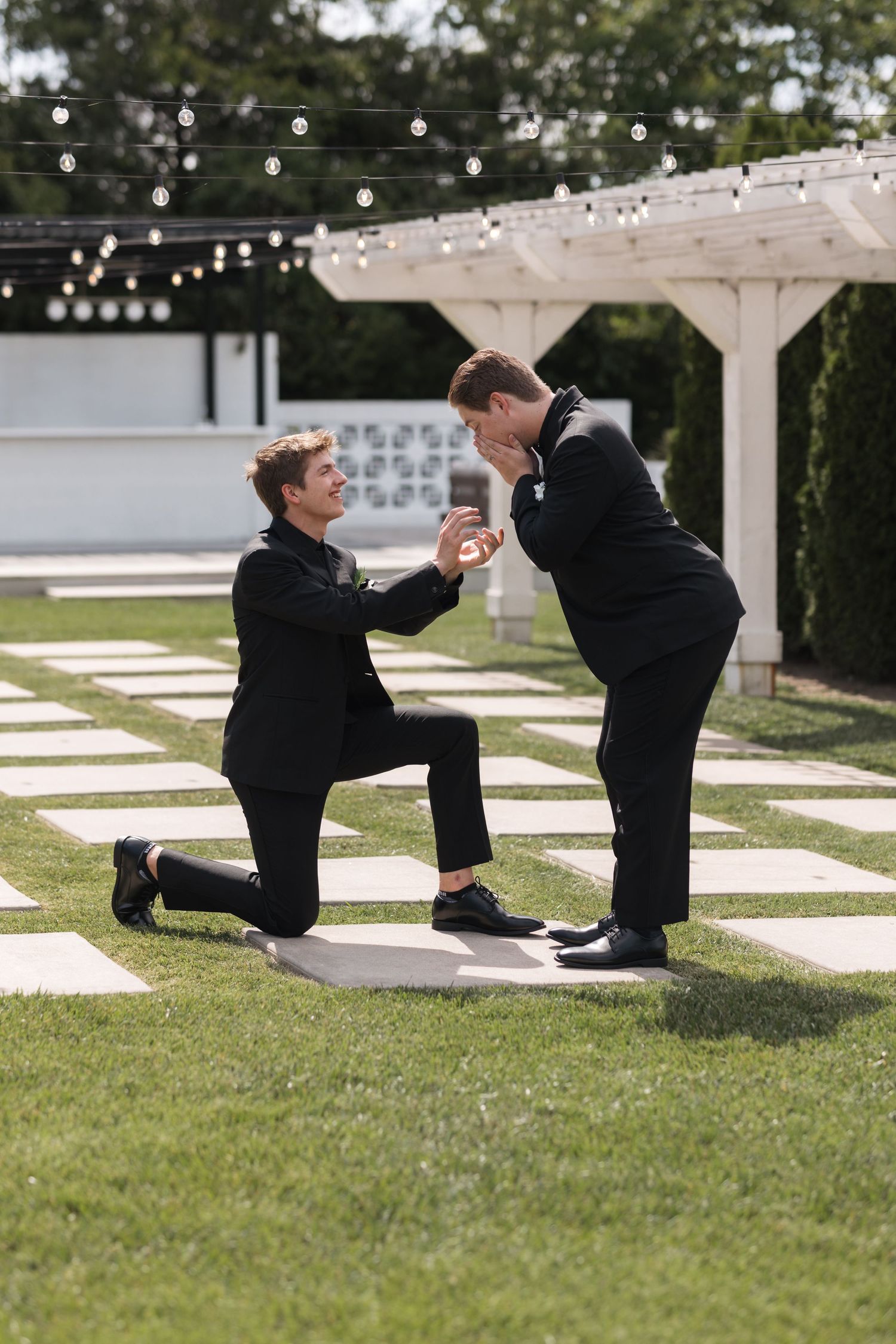 Two people in black suits share a special moment during an outdoor proposal under a pergola.