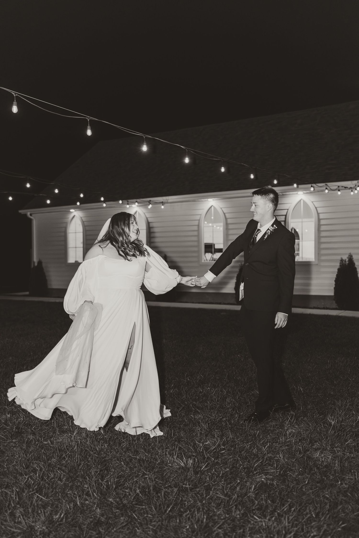 Black and white photo of couple dancing outside church with string lights overhead.