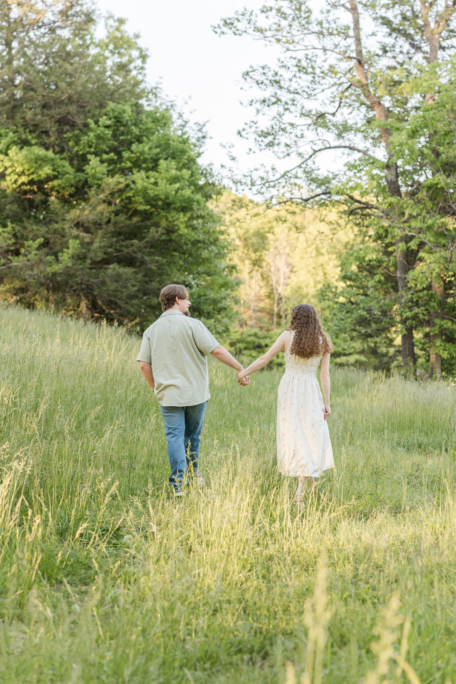 A couple walks hand in hand through a sun-lit meadow surrounded by tall grass and trees on a summer day.