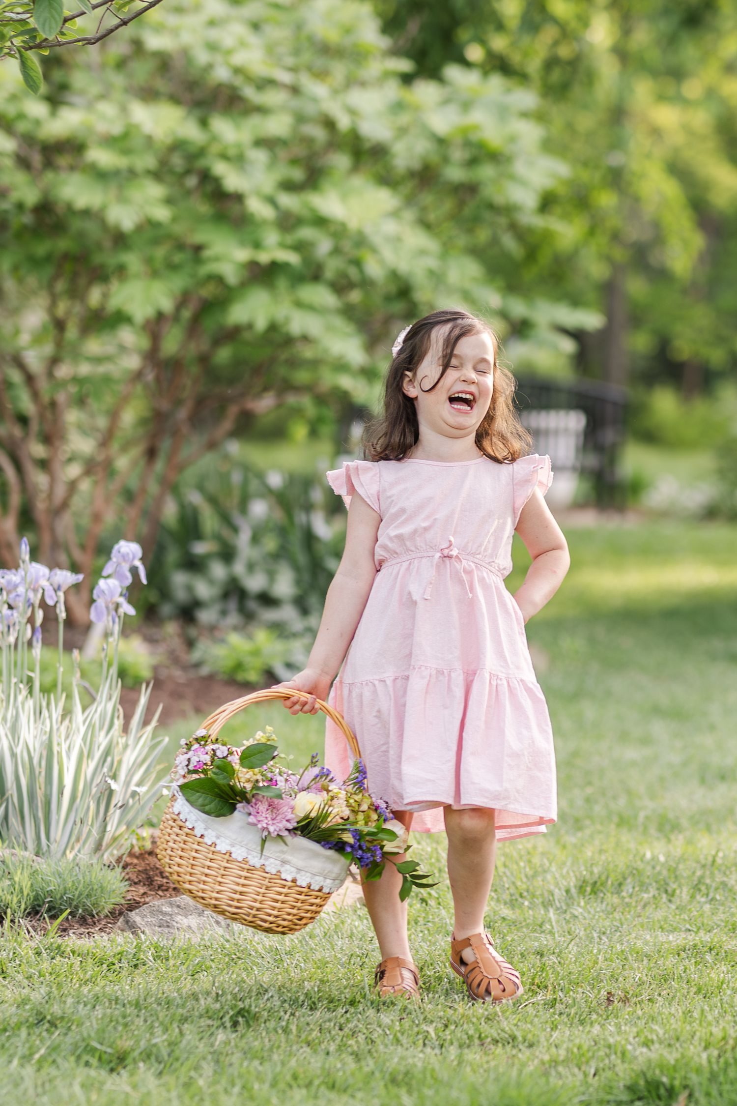 A child in a pink ruffled dress stands joyfully in a garden while holding a basket filled with flowers and plants.