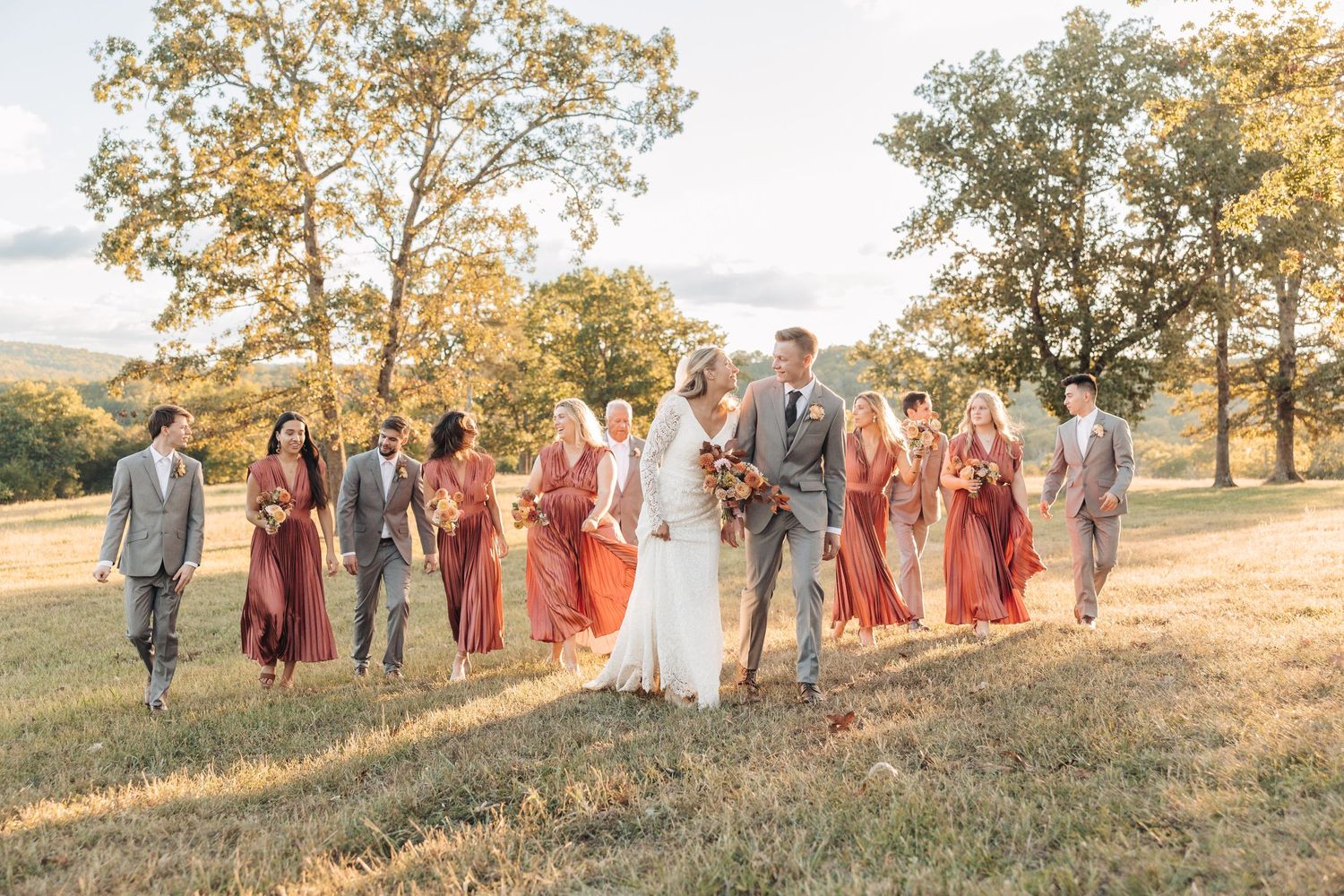 Wedding party in gray suits and burgundy dresses walking together on a hilltop during golden hour.