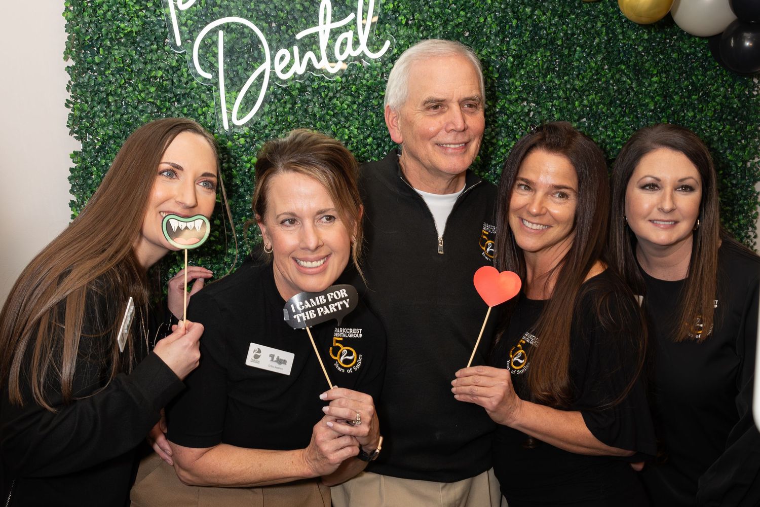 Group photo of dental staff members posing with prop signs against a green wall backdrop.
