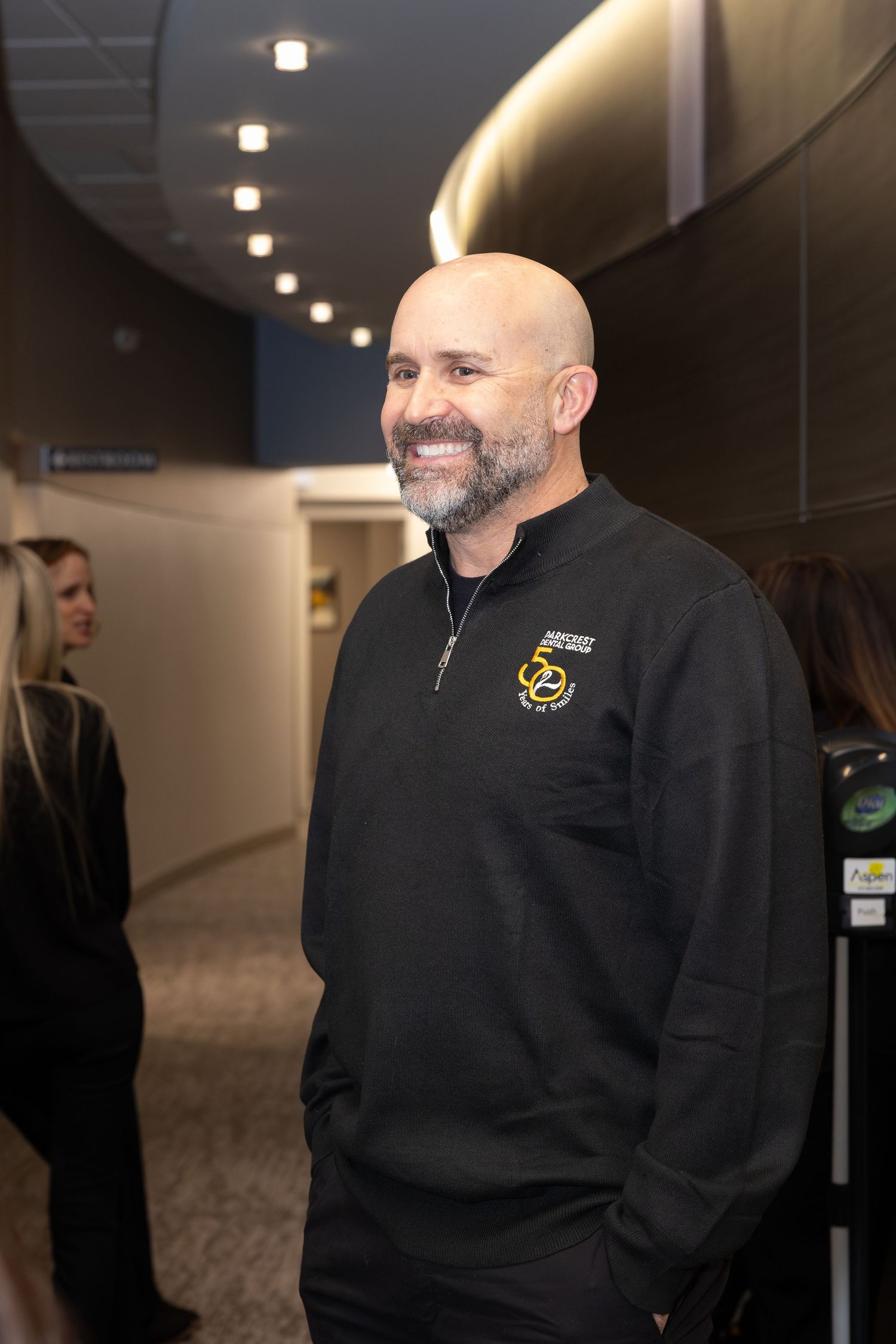 Person in black quarter-zip pullover with logo smiling in office hallway.