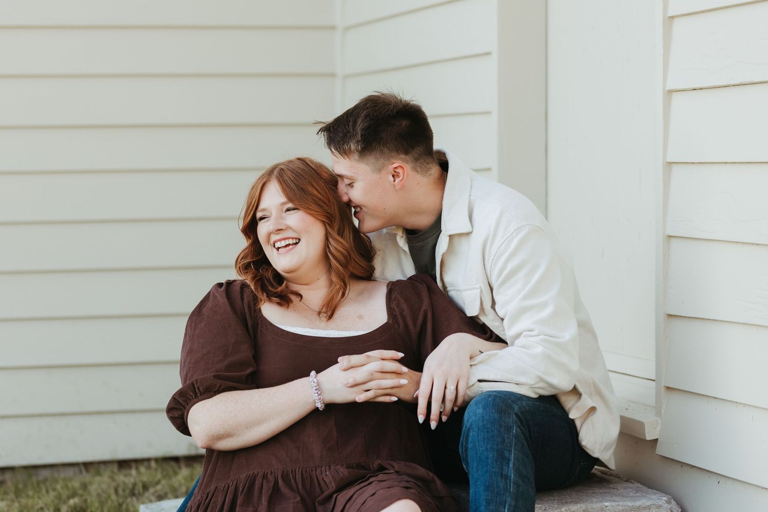 Intimate moment between couple sitting against white house wall.