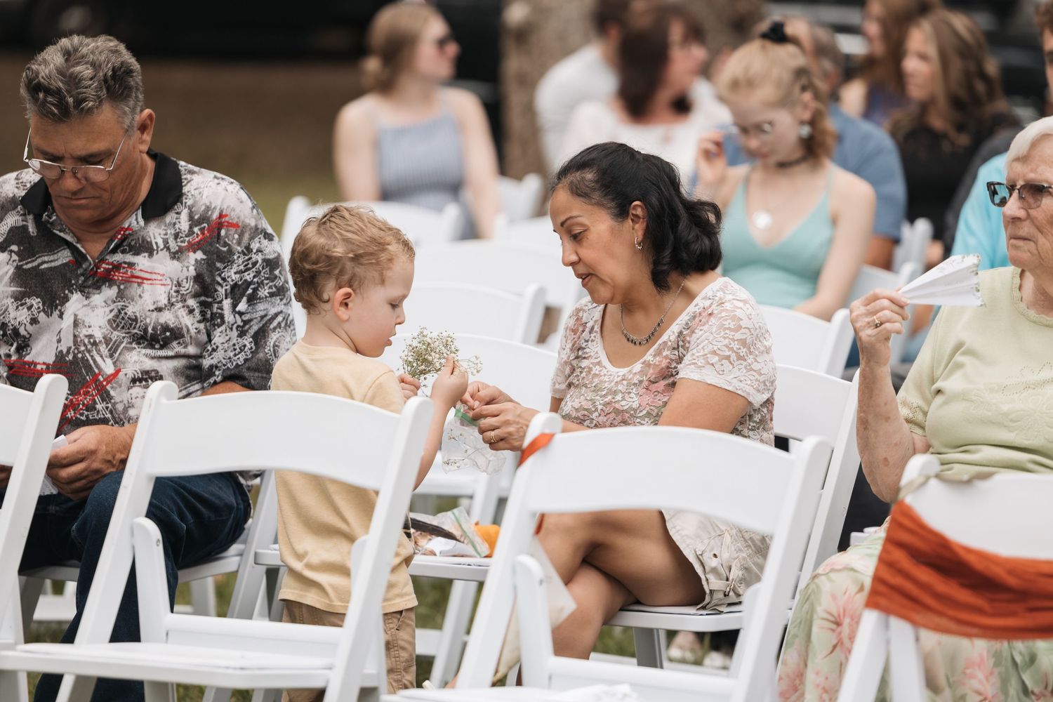 Guests seated in white chairs at an outdoor wedding ceremony interact during a casual moment.