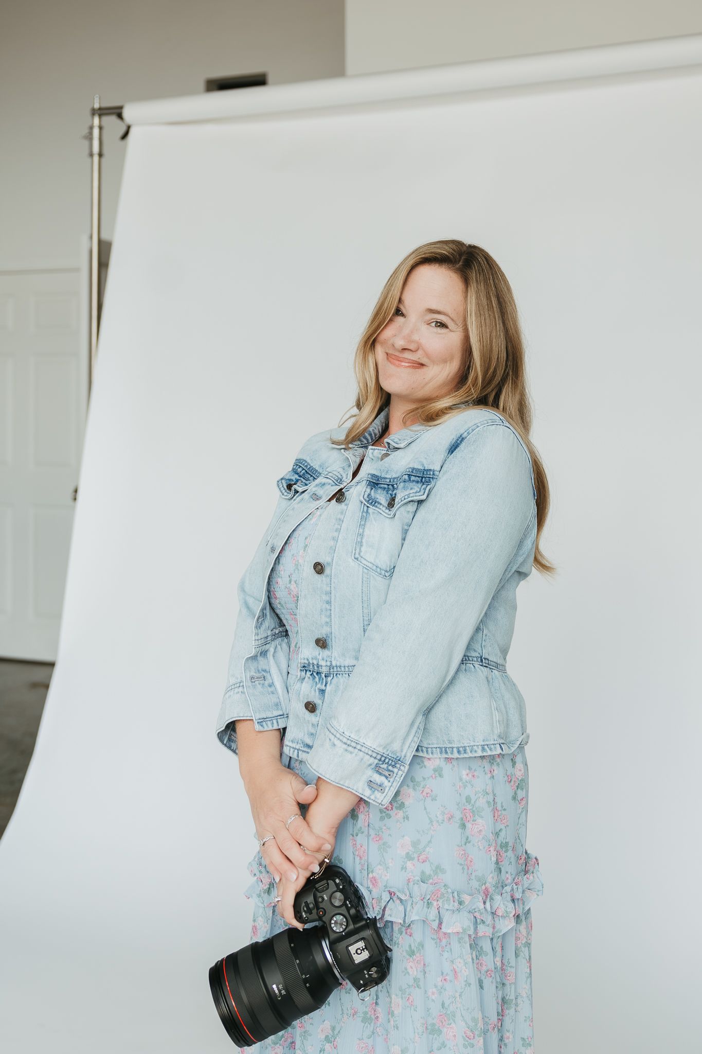 Professional fashion photoshoot showcasing person wearing light denim jacket over floral dress with camera in studio setting.
