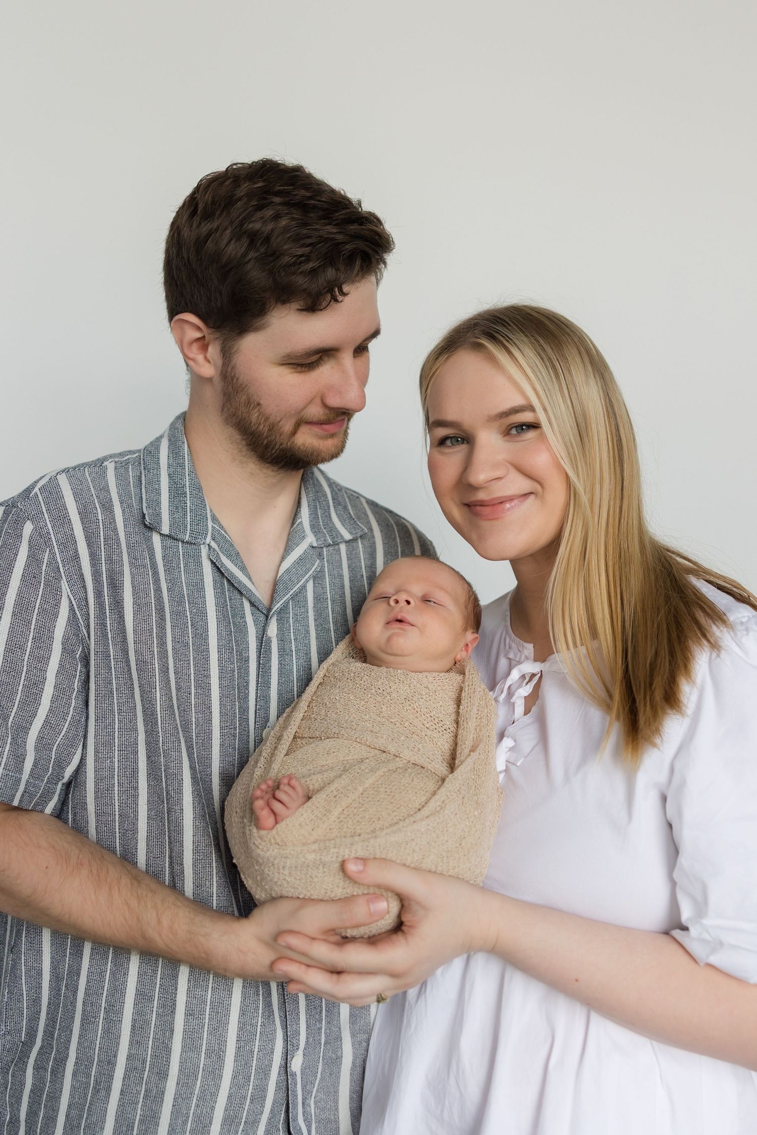 A couple shares loving glances while cradling their swaddled newborn baby in a minimalist studio setting.