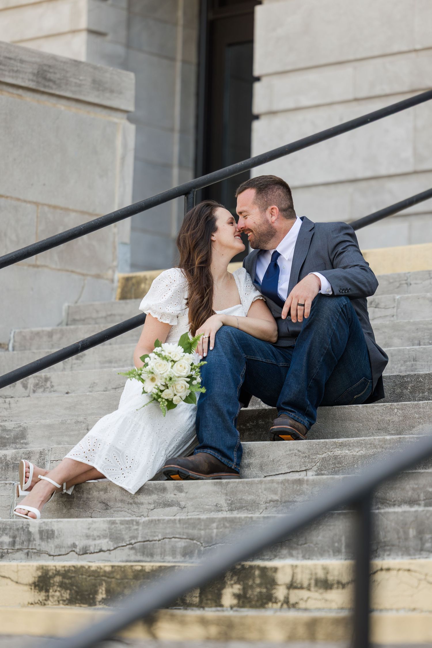 Romantic couple in formal attire sitting on stone steps with white floral bouquet and black railing.