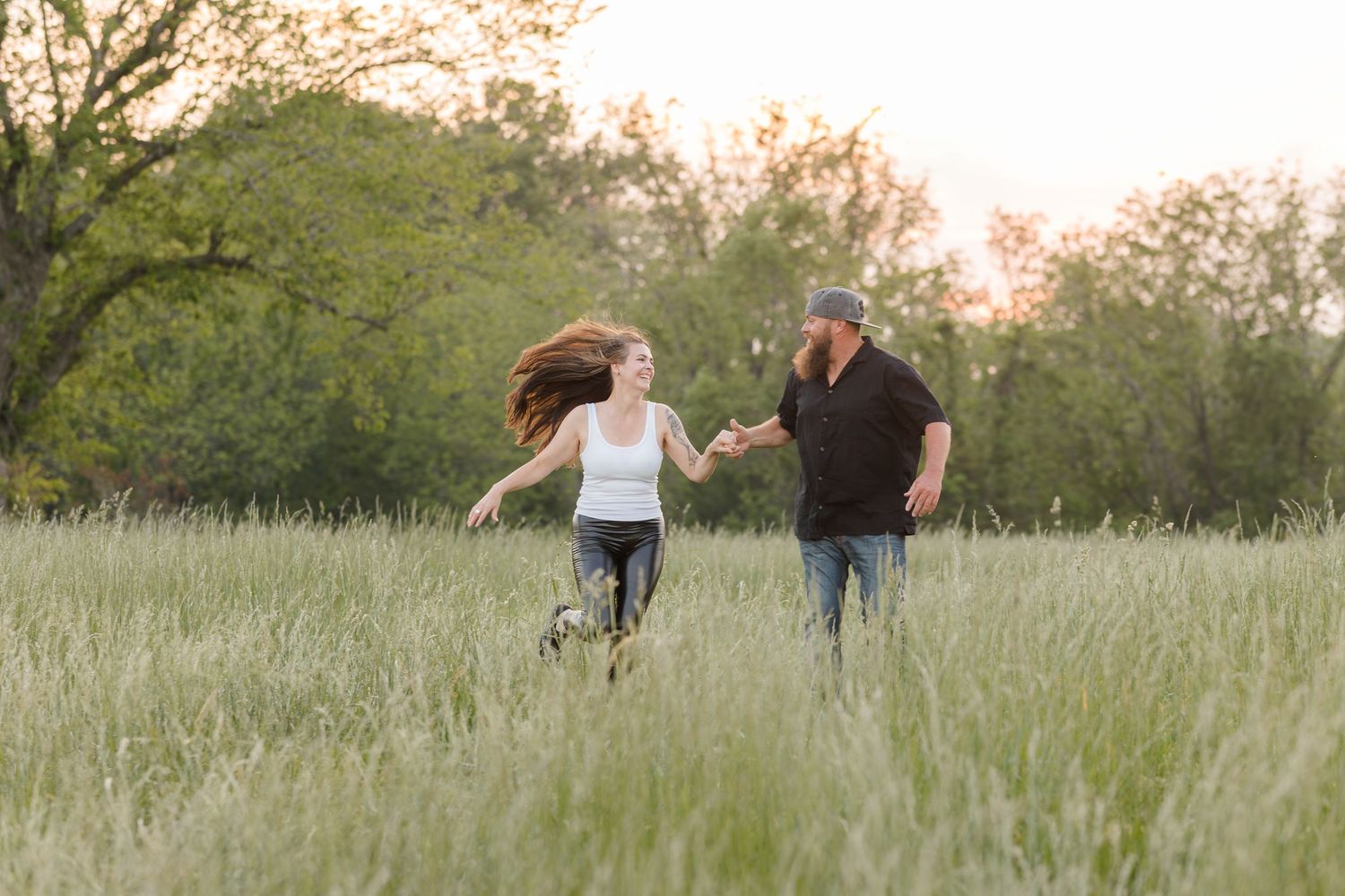 Two people run joyfully through a tall grass meadow at sunset with trees in the background.