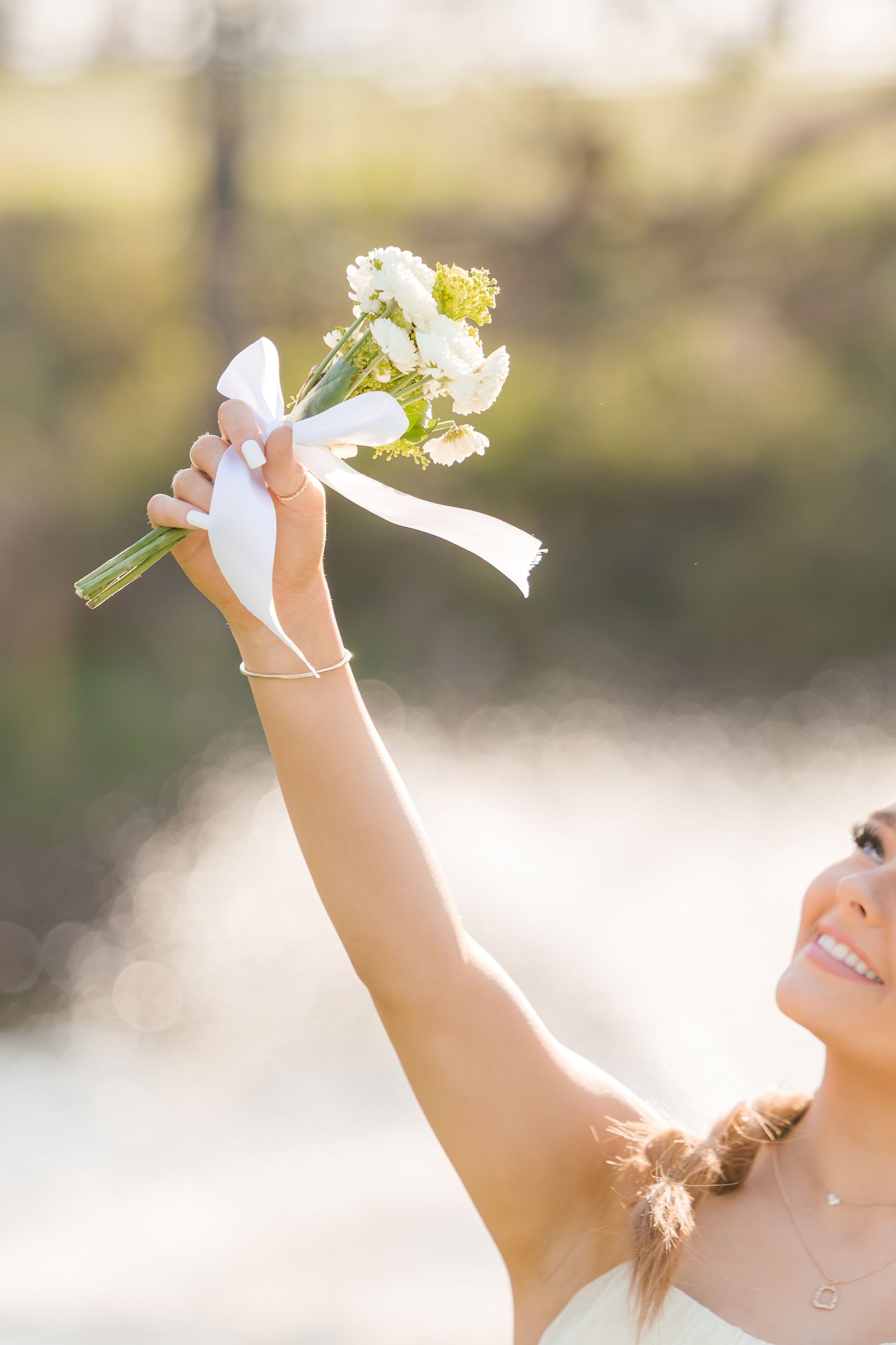 A joyful hand raises a delicate white and yellow bouquet with a ribbon against a soft sunlit background.