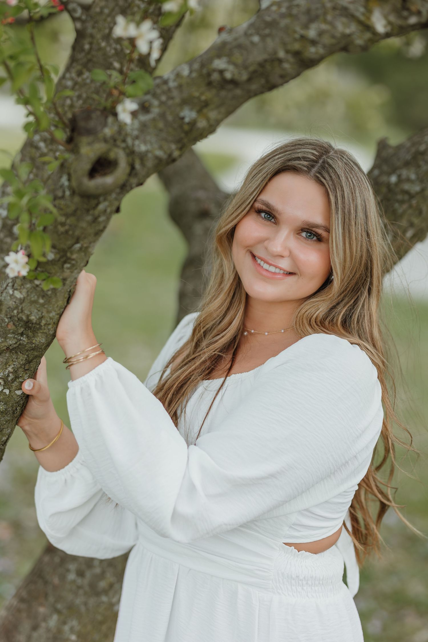 A person in a white summer dress poses next to a flowering tree branch in a natural outdoor setting.