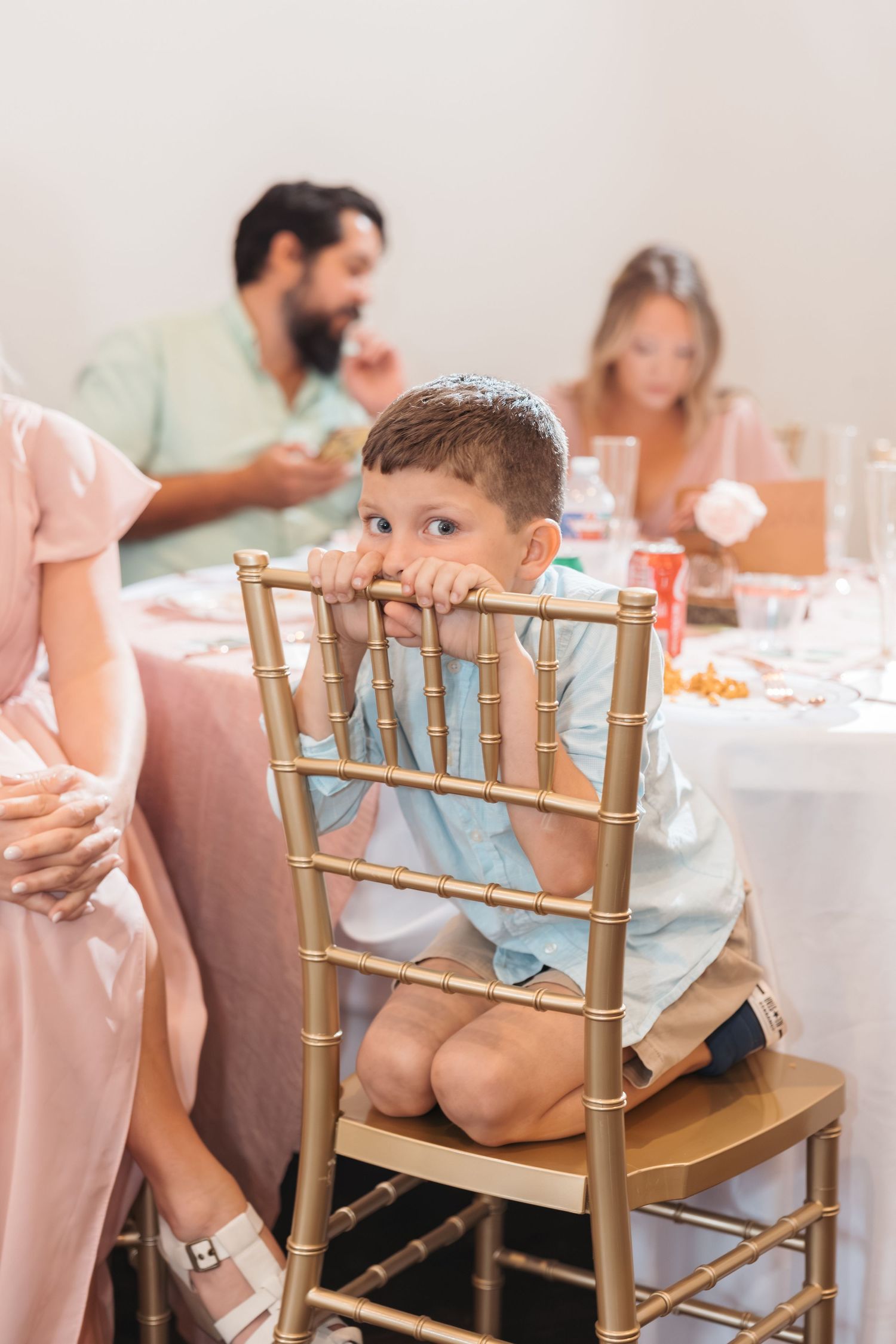 A young boy kneels on a gold chiavari chair at a family dinner party, resting his chin on the chair back.