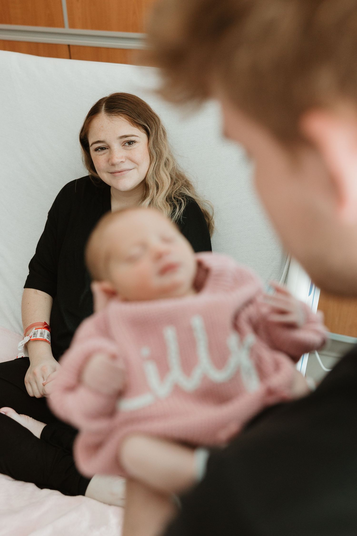 An infant in a pink sweater rests on a white surface while others gather around in a cozy indoor setting.