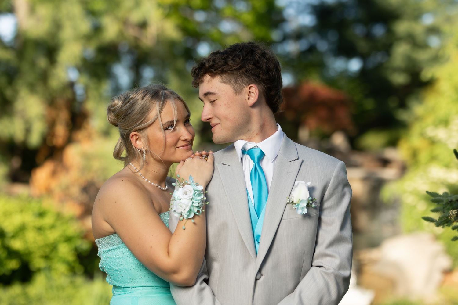 Young couple in prom attire, girl in teal dress with wrist corsage, boy in gray suit with turquoise tie, outdoors.