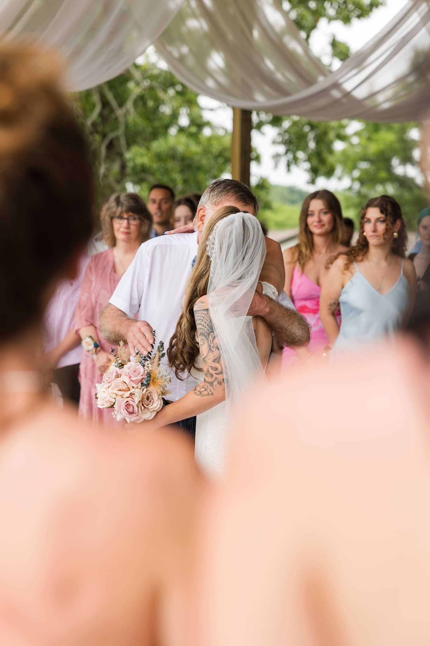 Bride and groom share a first kiss at outdoor wedding ceremony with bridesmaids in pink and blue dresses.