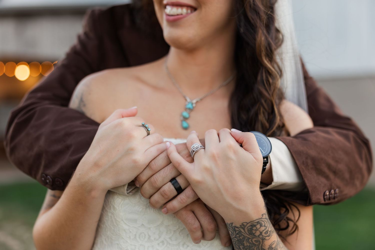 Bride in lace dress with turquoise necklace and diamond ring, embraced by groom in brown jacket.
