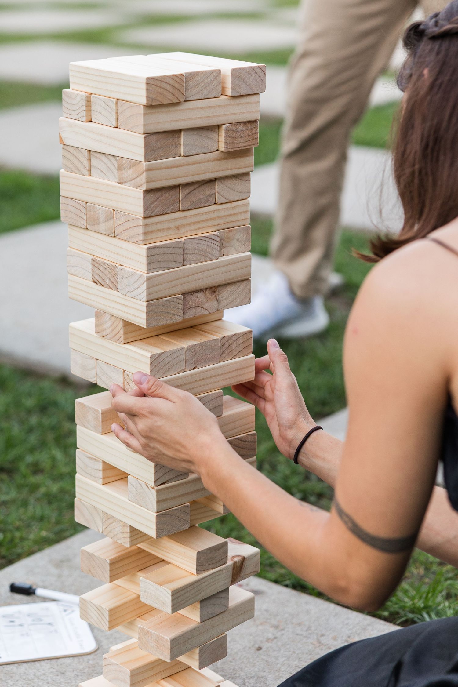 Woman carefully removing a wooden block from a tall Jenga tower during an outdoor garden party.
