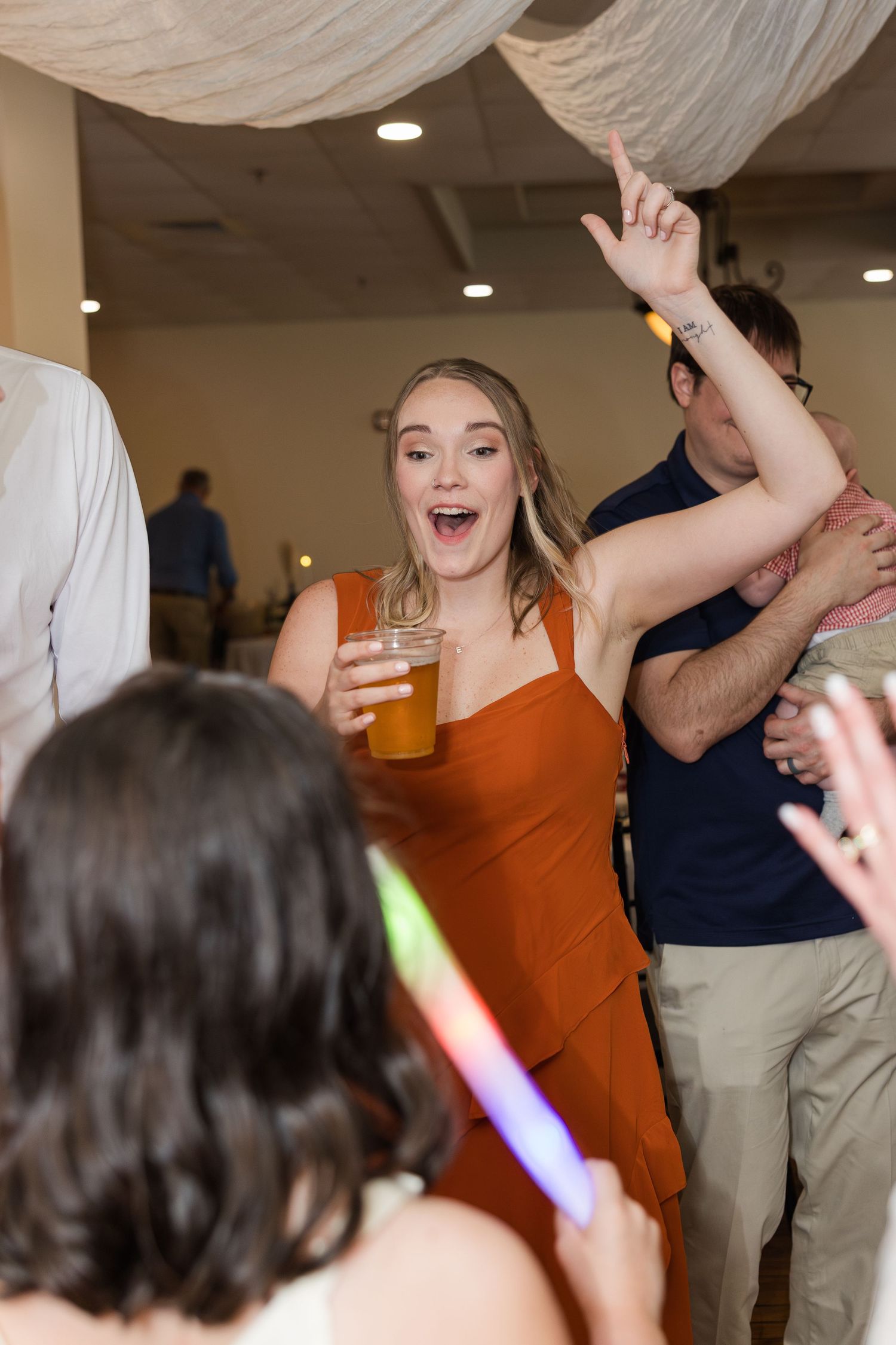 Excited wedding guest in orange dress dancing with a beer at a lively indoor wedding reception.