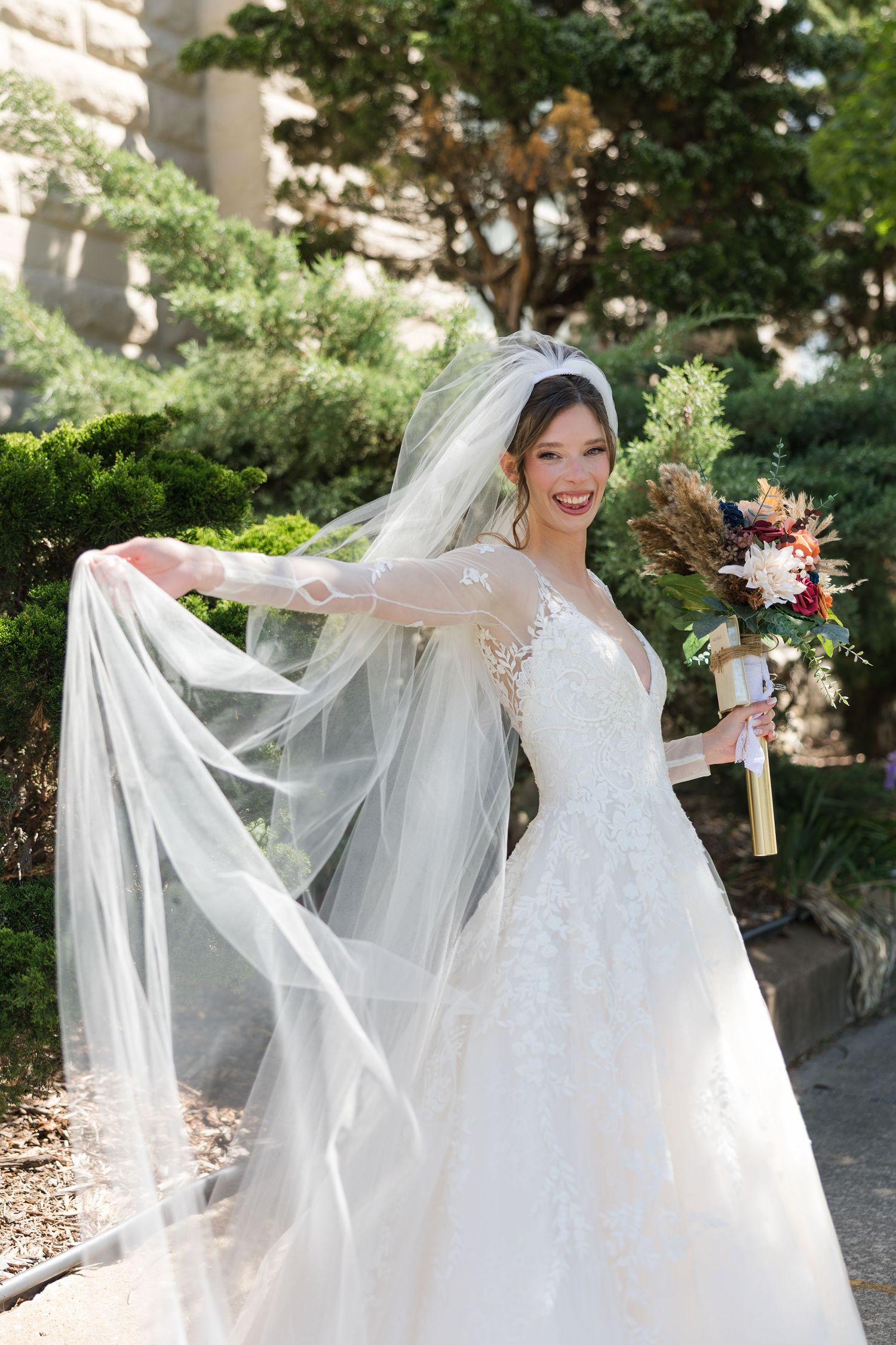 Smiling bride in a lace long-sleeve gown twirls her flowing veil while holding a bouquet outdoors.