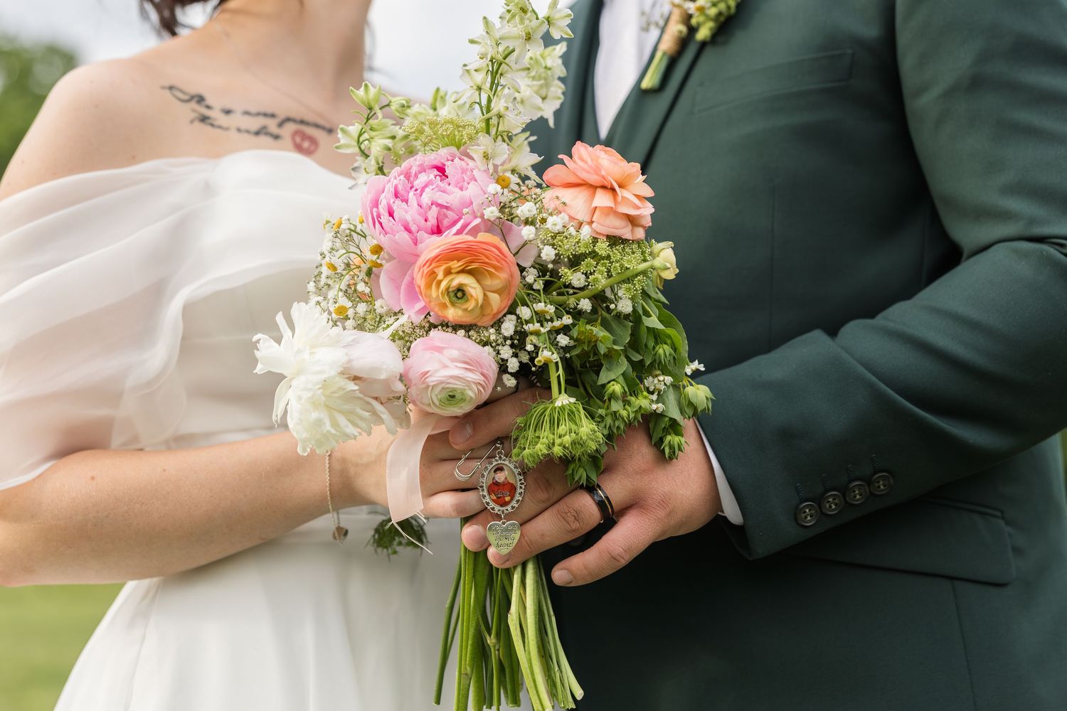 Bride and groom holding a colorful wedding bouquet with pink peonies, ranunculus, and greenery at an outdoor ceremony.