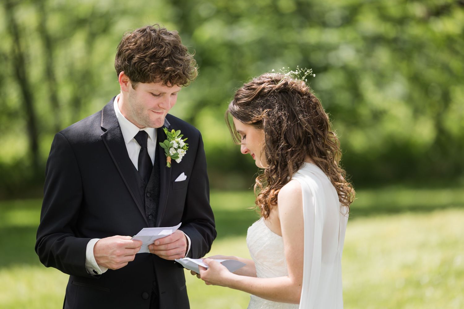 Bride and groom reading wedding vows from cards outdoors, surrounded by lush green trees on their wedding day.