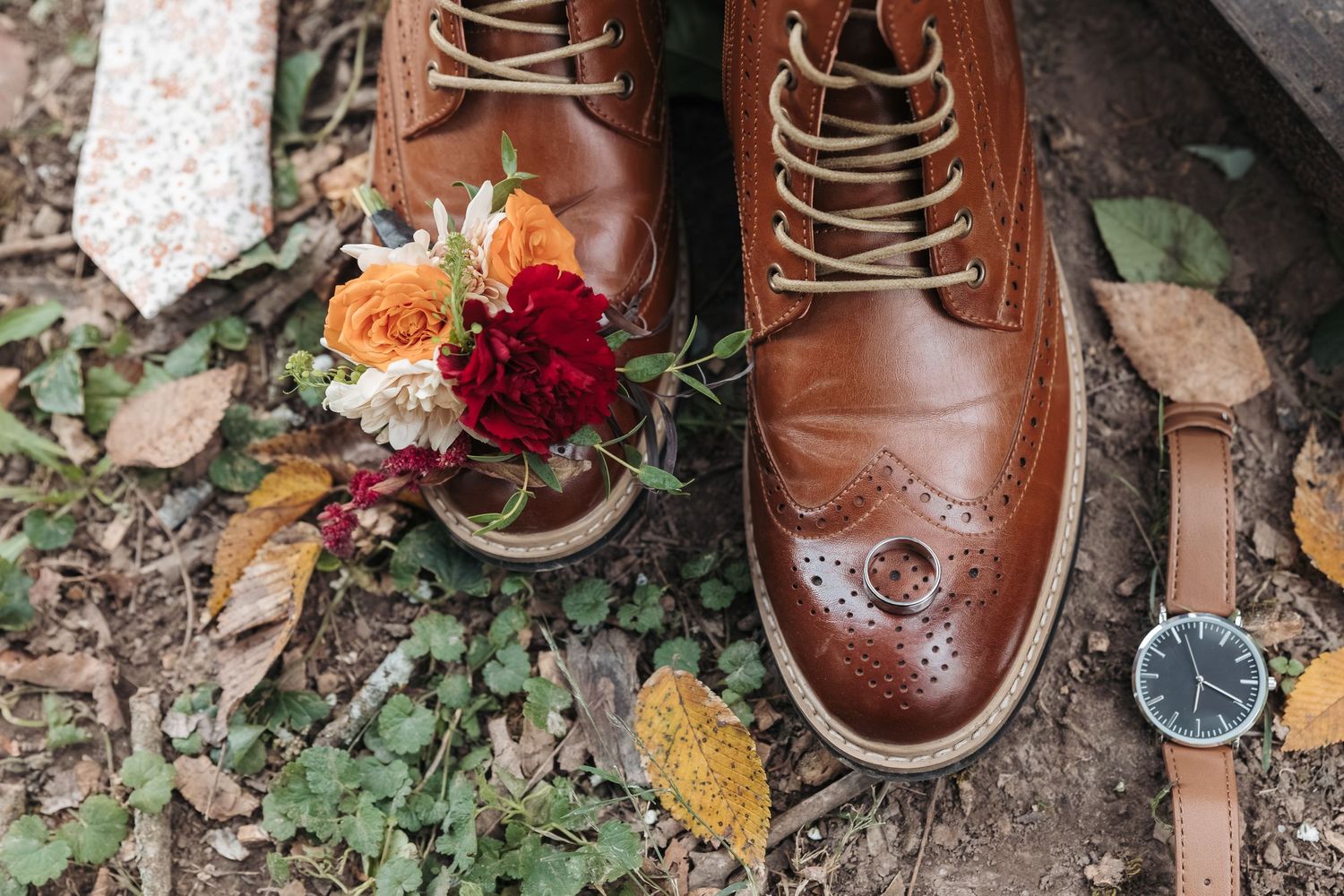 Brown leather dress shoes with autumn flowers and wristwatch on fallen leaves.
