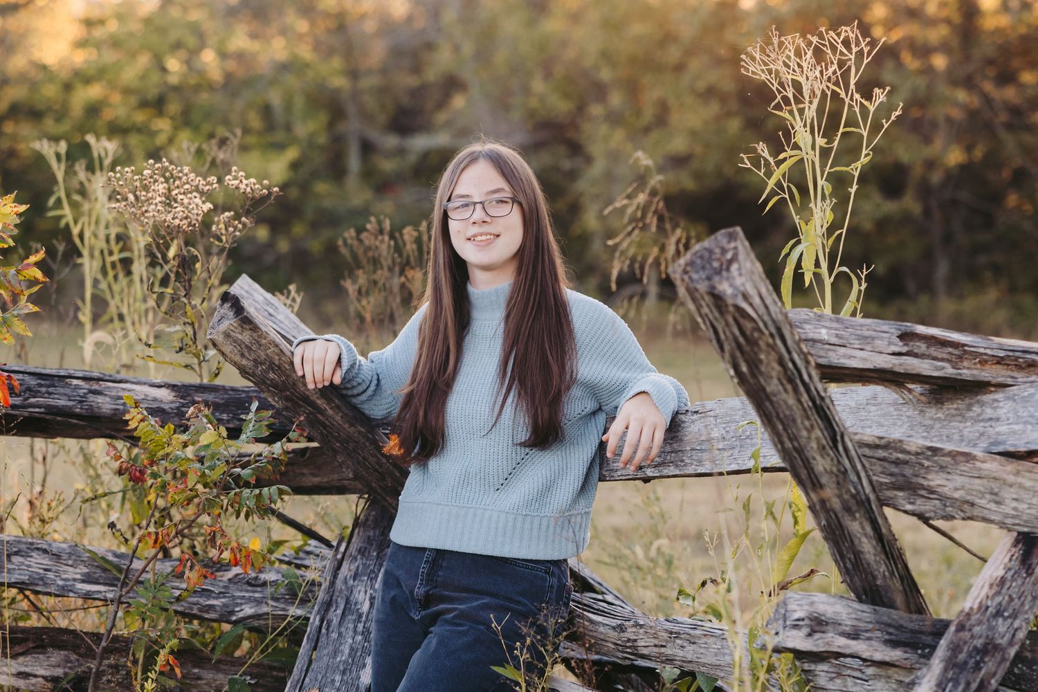 Young person leaning on rustic wooden fence with autumn foliage in background.