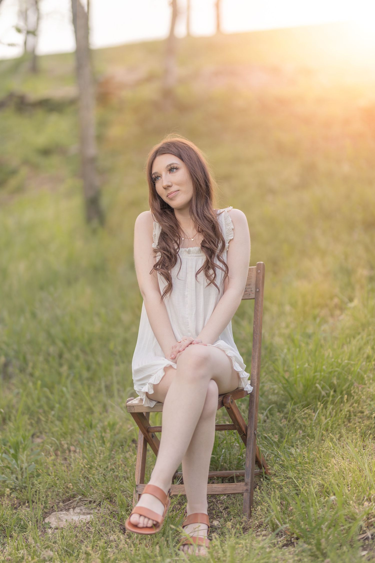 Woman in white sundress sits gracefully on wooden chair during outdoor sunset photoshoot.