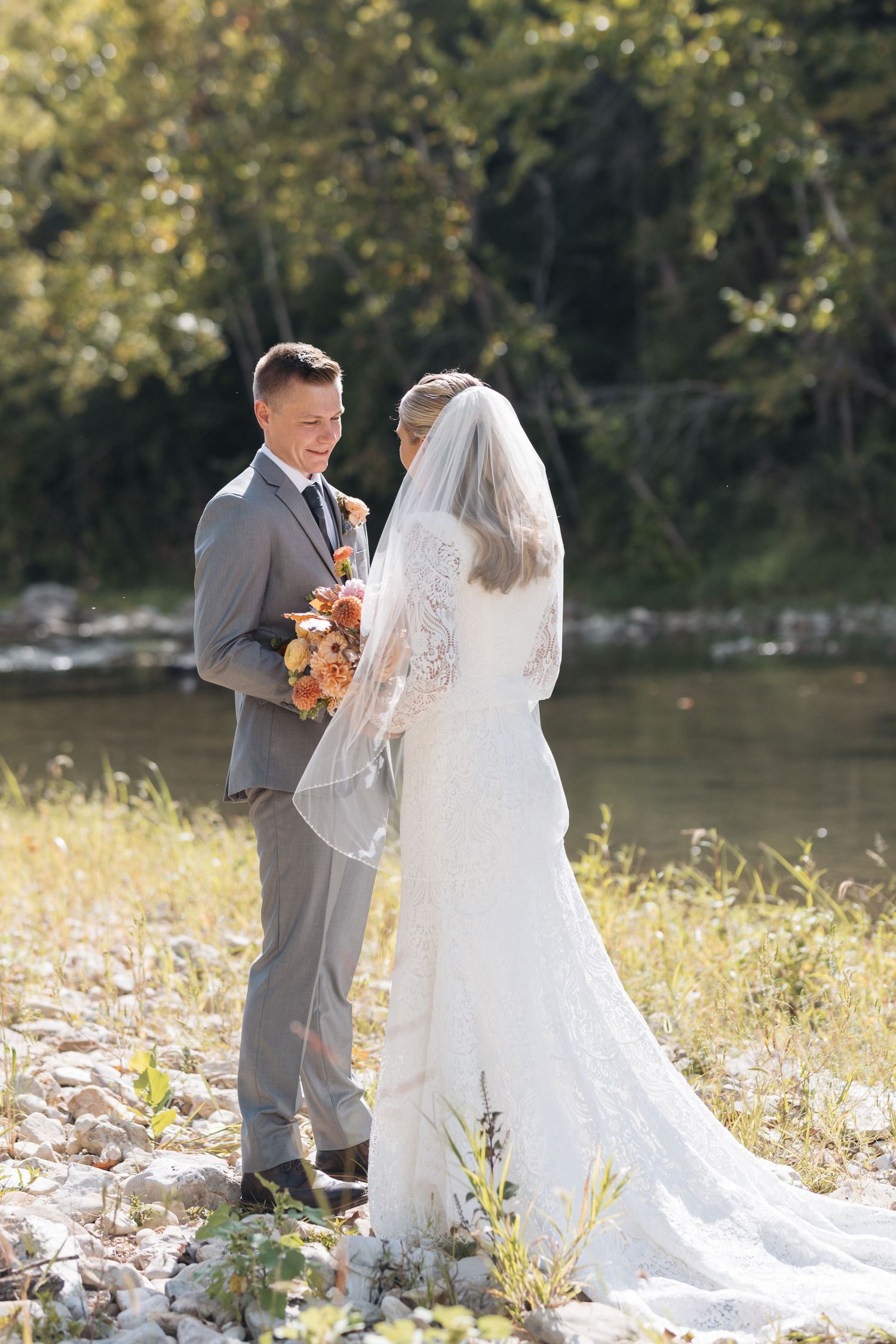 Bride and groom share intimate moment by riverside in wedding attire.